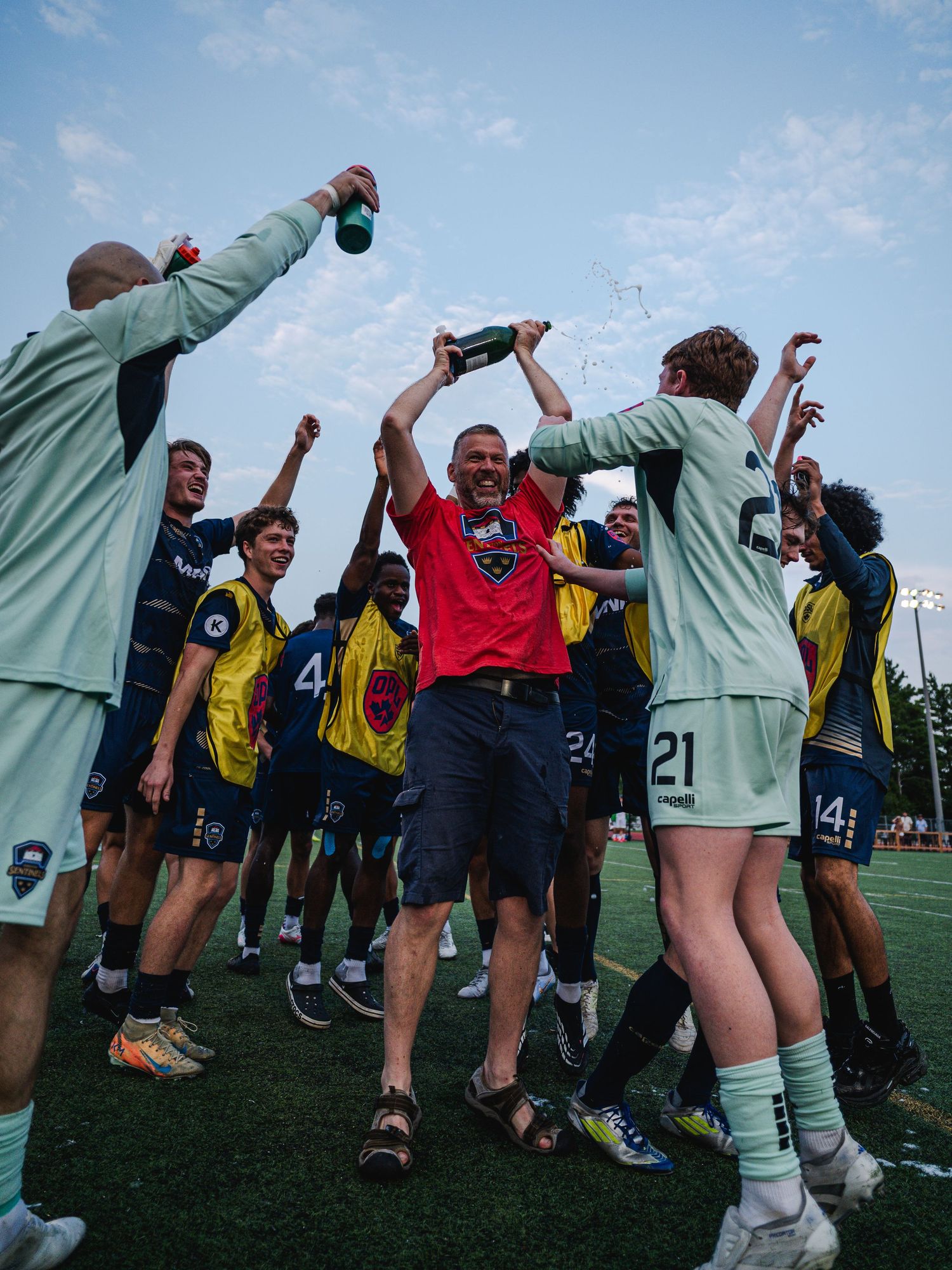Soccer team celebrates victory with coach in red shirt raising trophy amid cheering players on turf field.