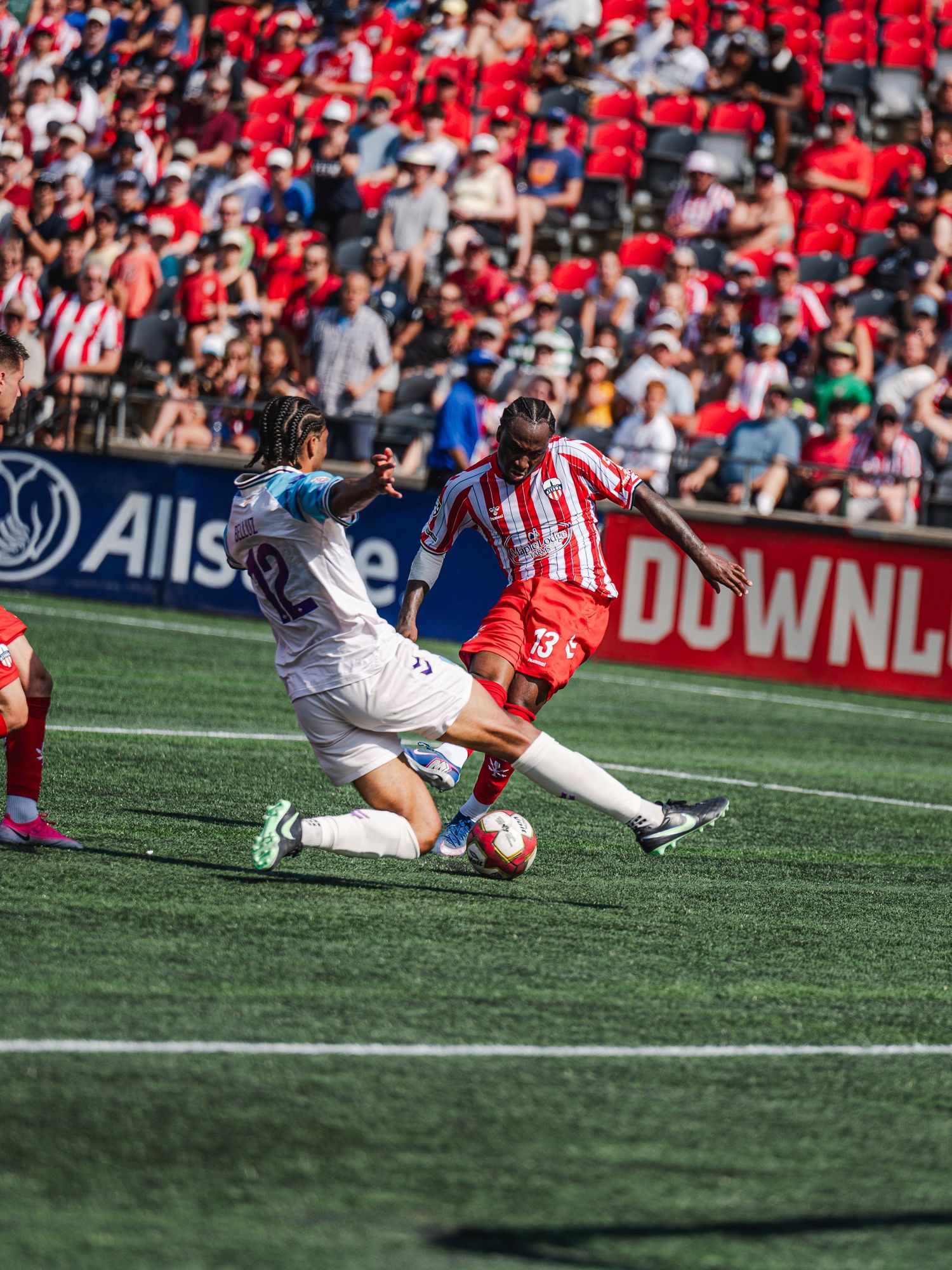 Soccer player in red jersey shoots ball while defender slides in during stadium match.