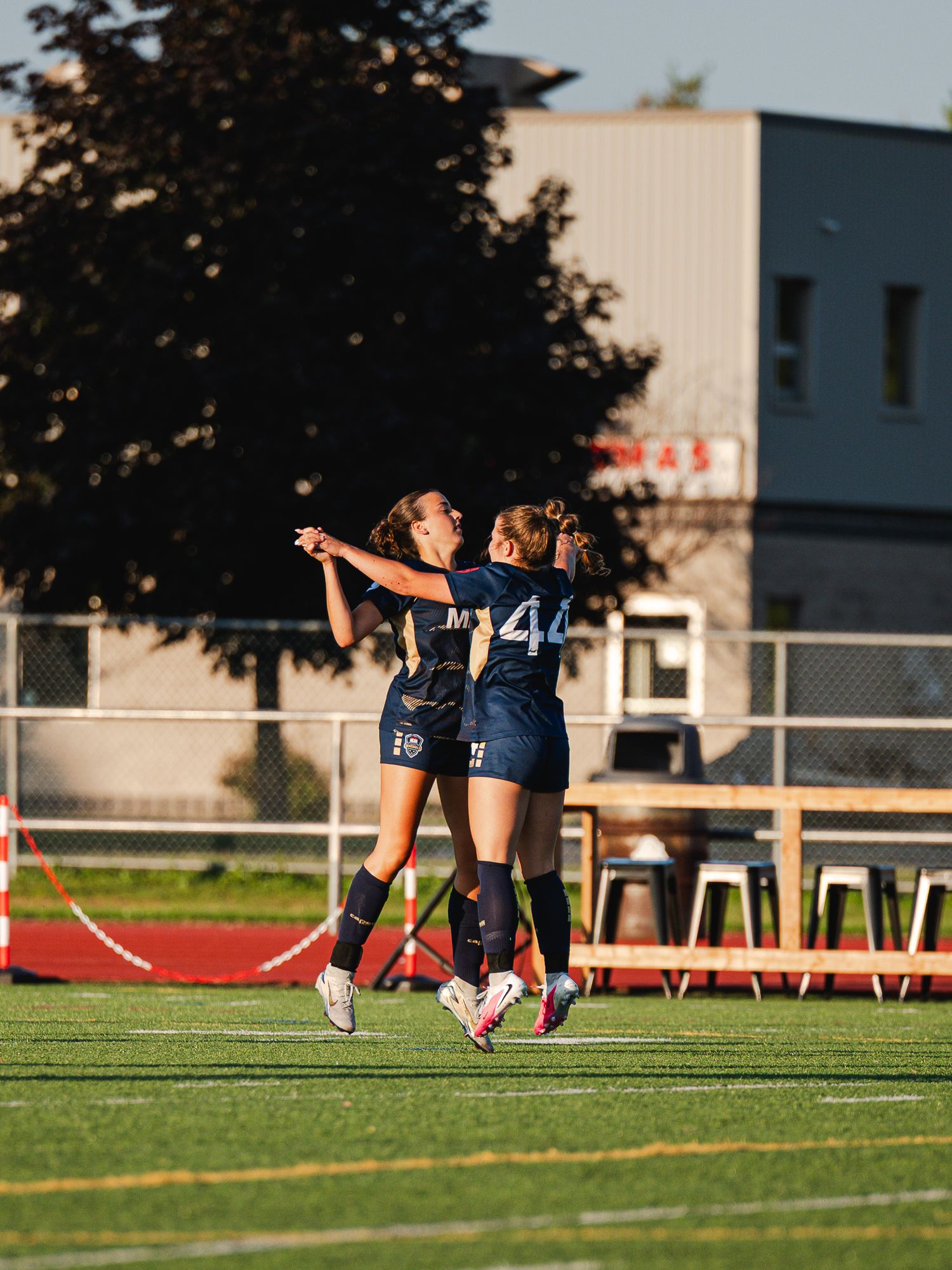 Two female soccer players in navy uniforms celebrate together on a sunny green field after scoring a goal.