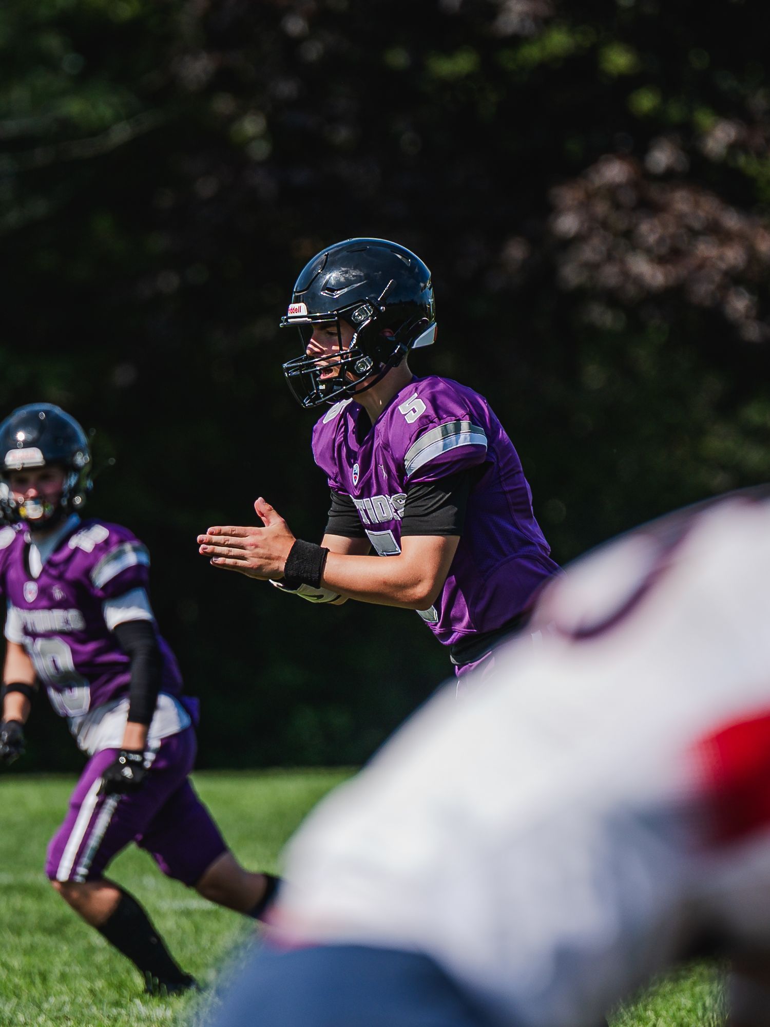 Quarterback in purple uniform claps hands at the line of scrimmage during a sunny outdoor football game.