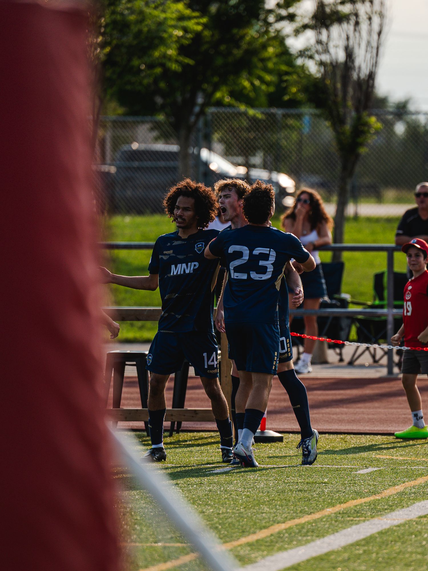 Two MNP soccer players in navy jerseys celebrate together on the sideline after a goal.
