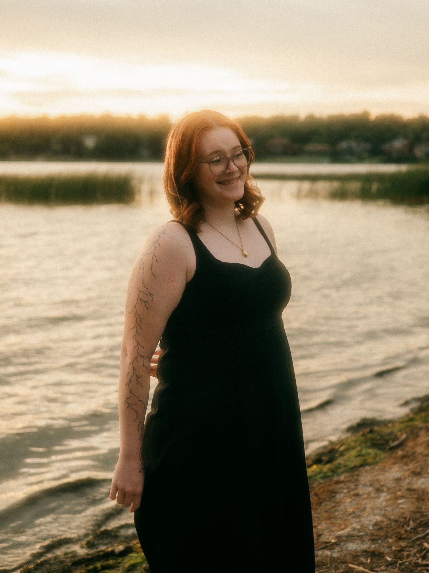 Smiling tattooed woman in black dress standing by lake at golden sunset hour.