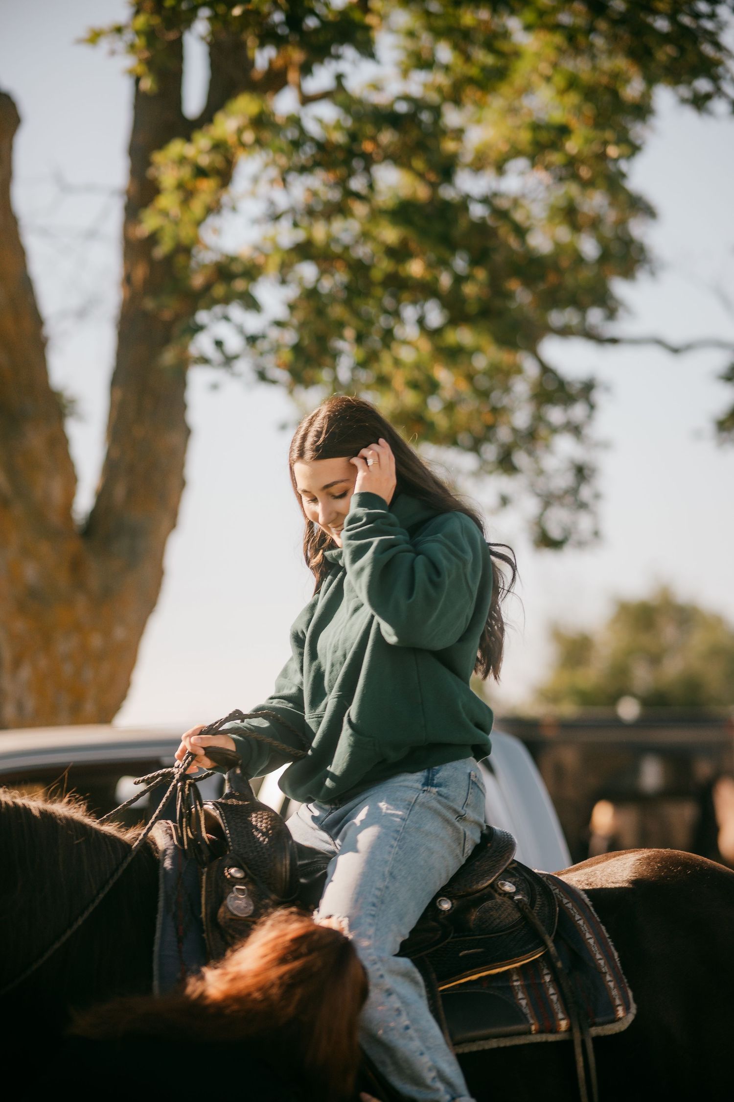 Young woman riding dark horse outdoors beneath large tree in sunlight.