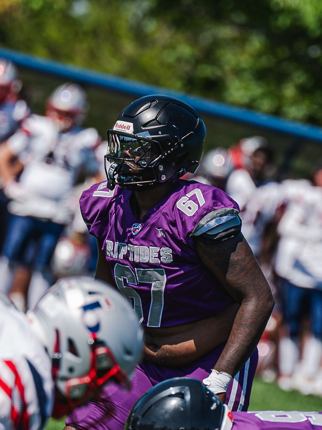 Football player number 67 in purple Riptides jersey and black helmet stands ready during a game.