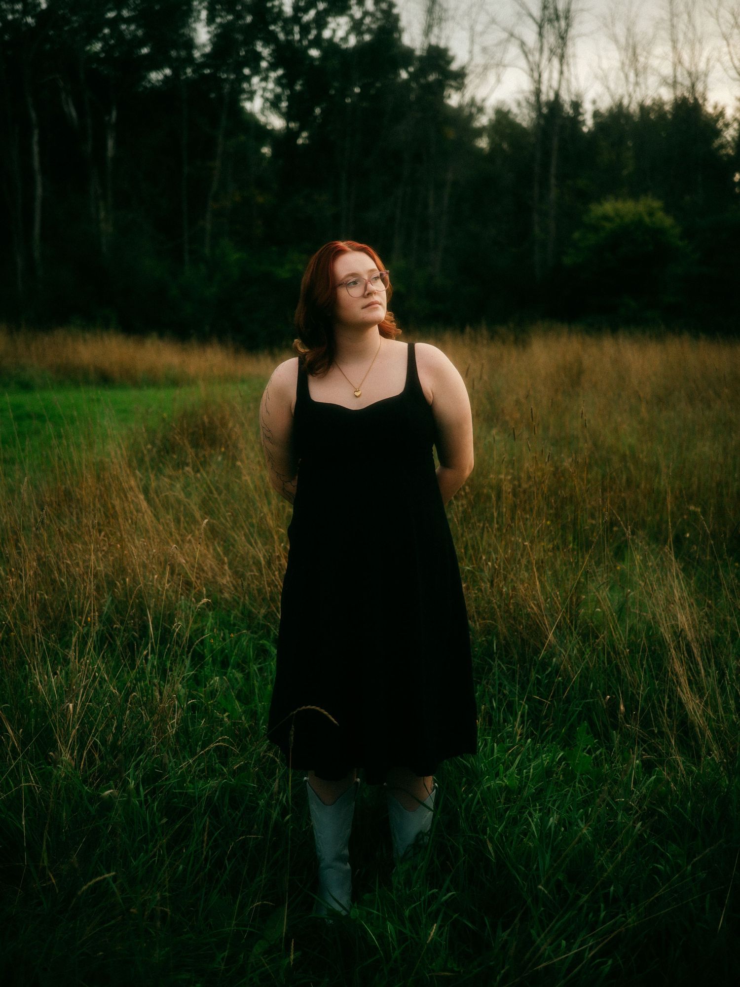 Young woman with red hair in black dress standing in tall grass field at dusk near forest.