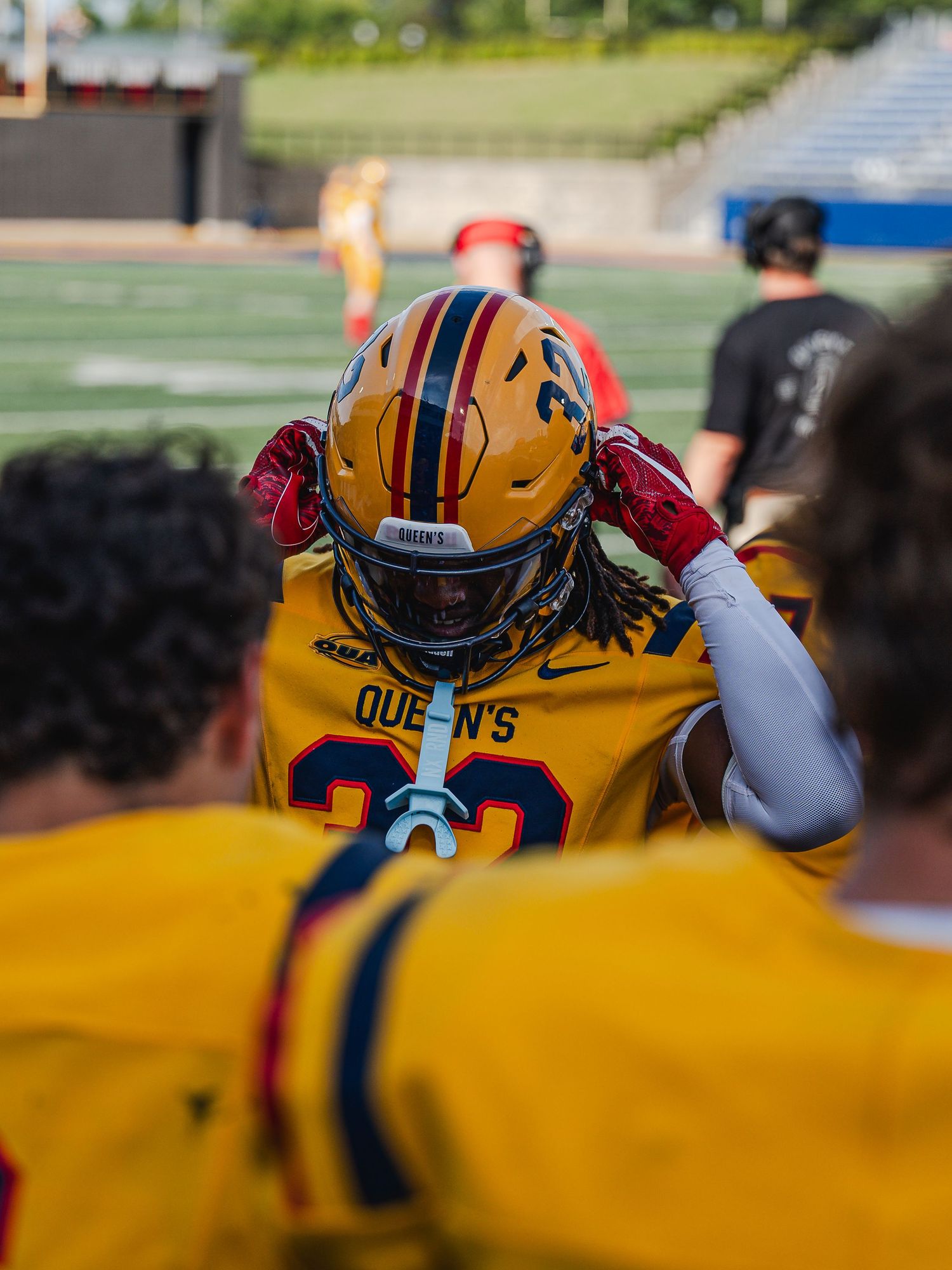 Queen's football player #30 adjusts red helmet during team huddle on the sideline.