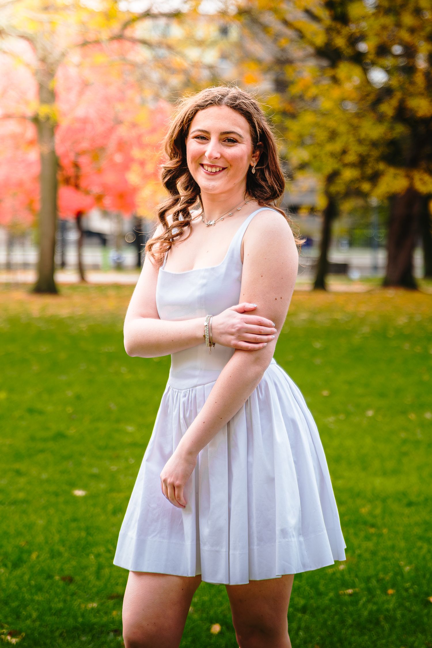 Smiling young woman in white dress stands in park with colorful autumn foliage in background.