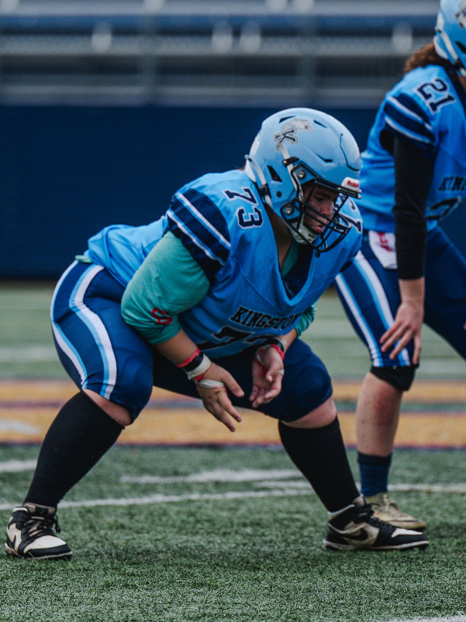 Football player #73 in blue Kingston uniform crouches in defensive stance on field.