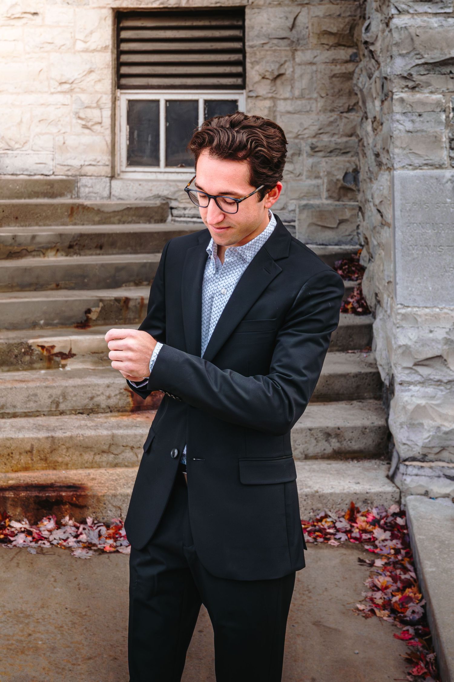 Well-dressed young man in black suit adjusts cufflinks outside stone university building.