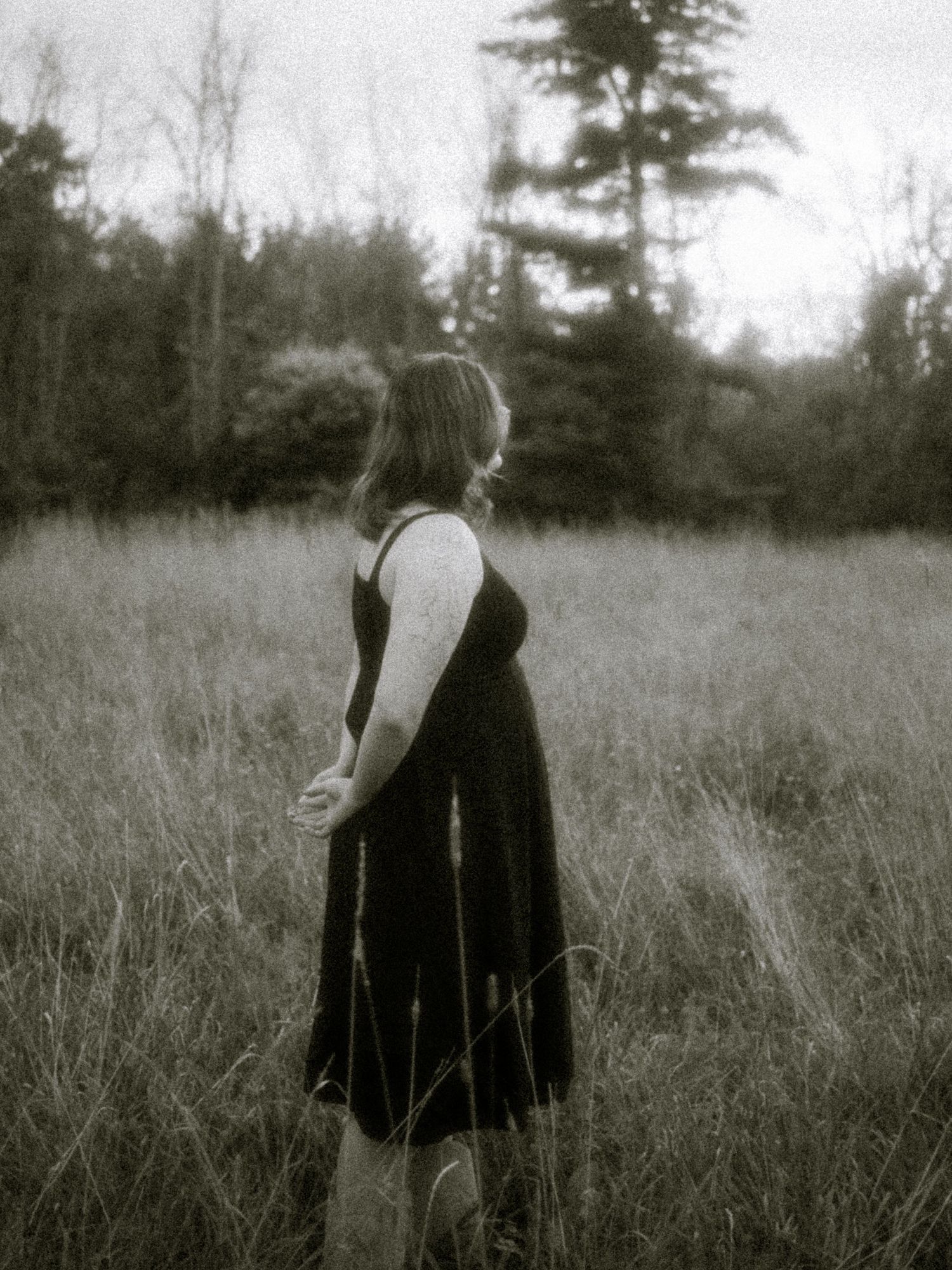A woman in a black dress stands in a grassy field surrounded by trees in a vintage black and white photo.