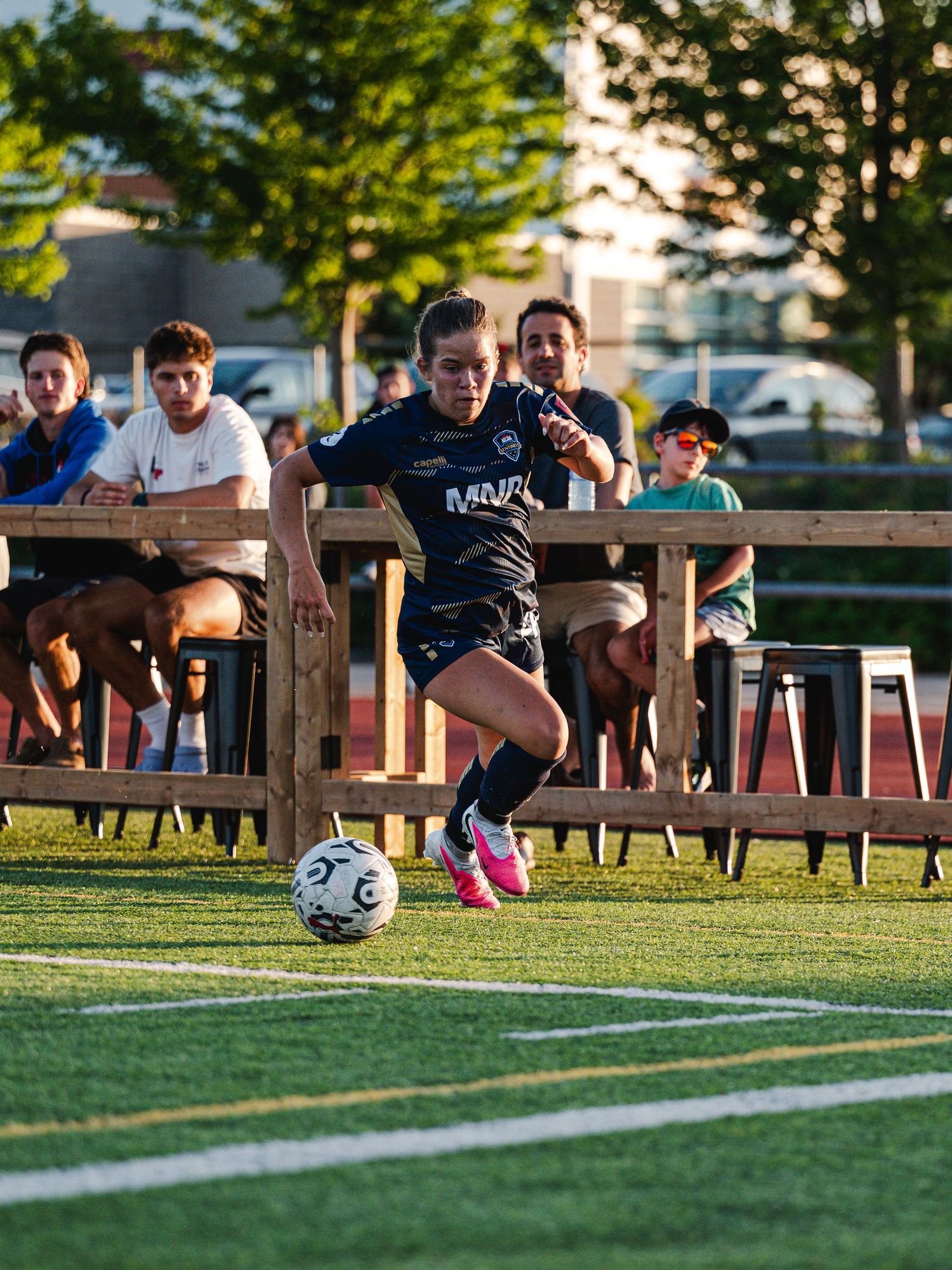 Female soccer player in black uniform dribbles the ball near the sideline with spectators watching from bleachers.