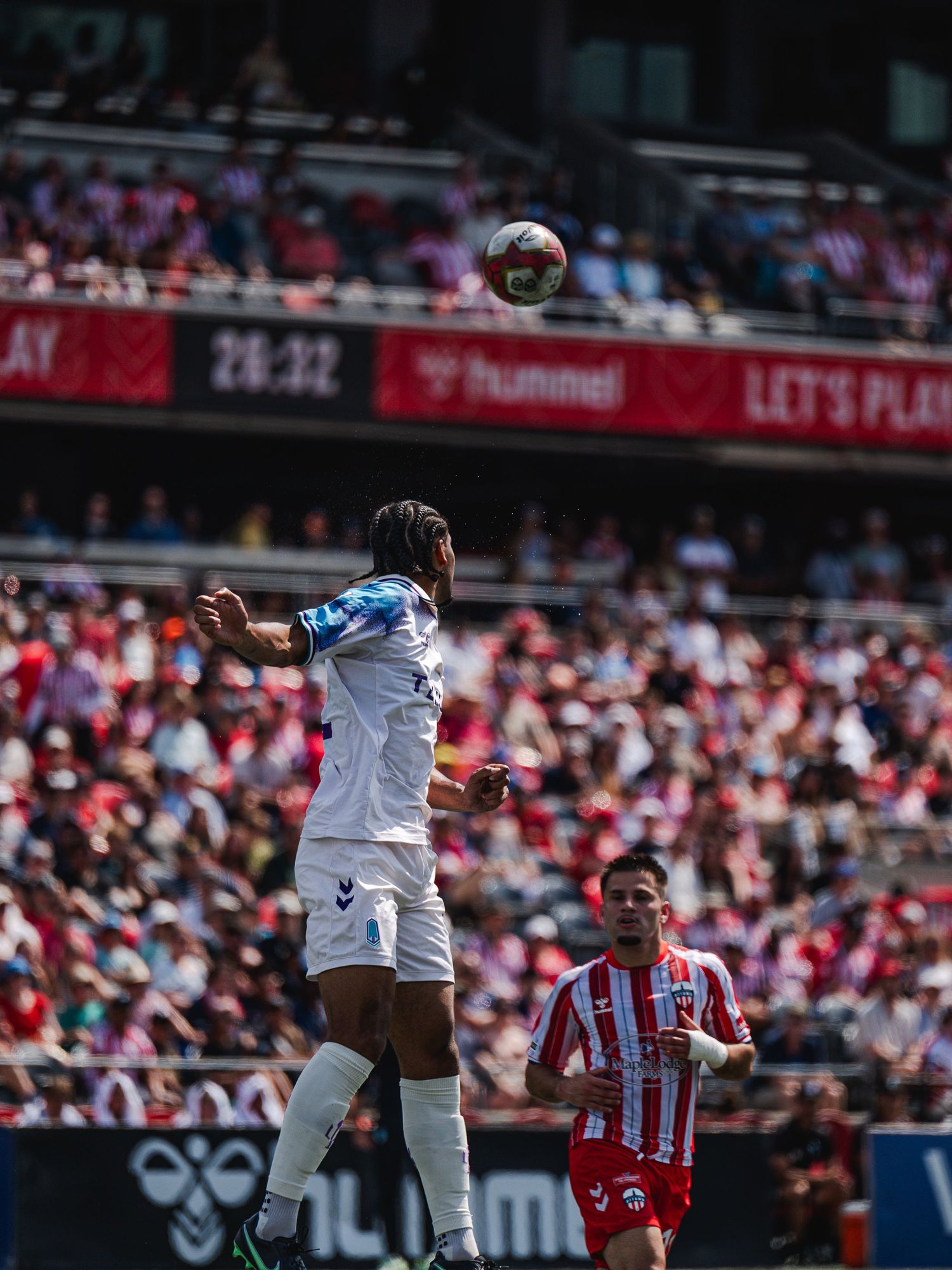 Soccer player in white jersey wins aerial header duel during packed stadium match.