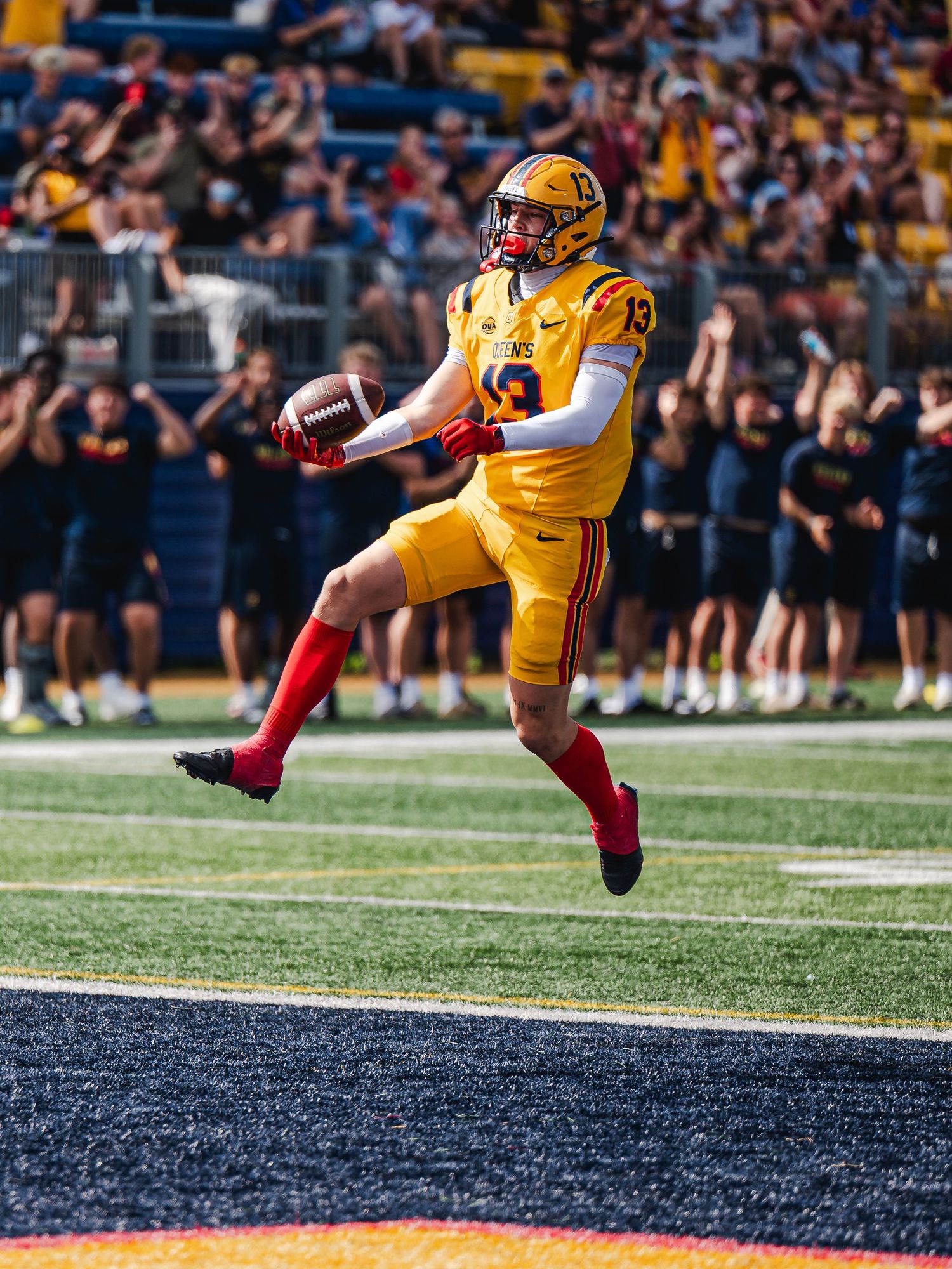 Queen's football player #13 in gold uniform leaps triumphantly while scoring a touchdown.