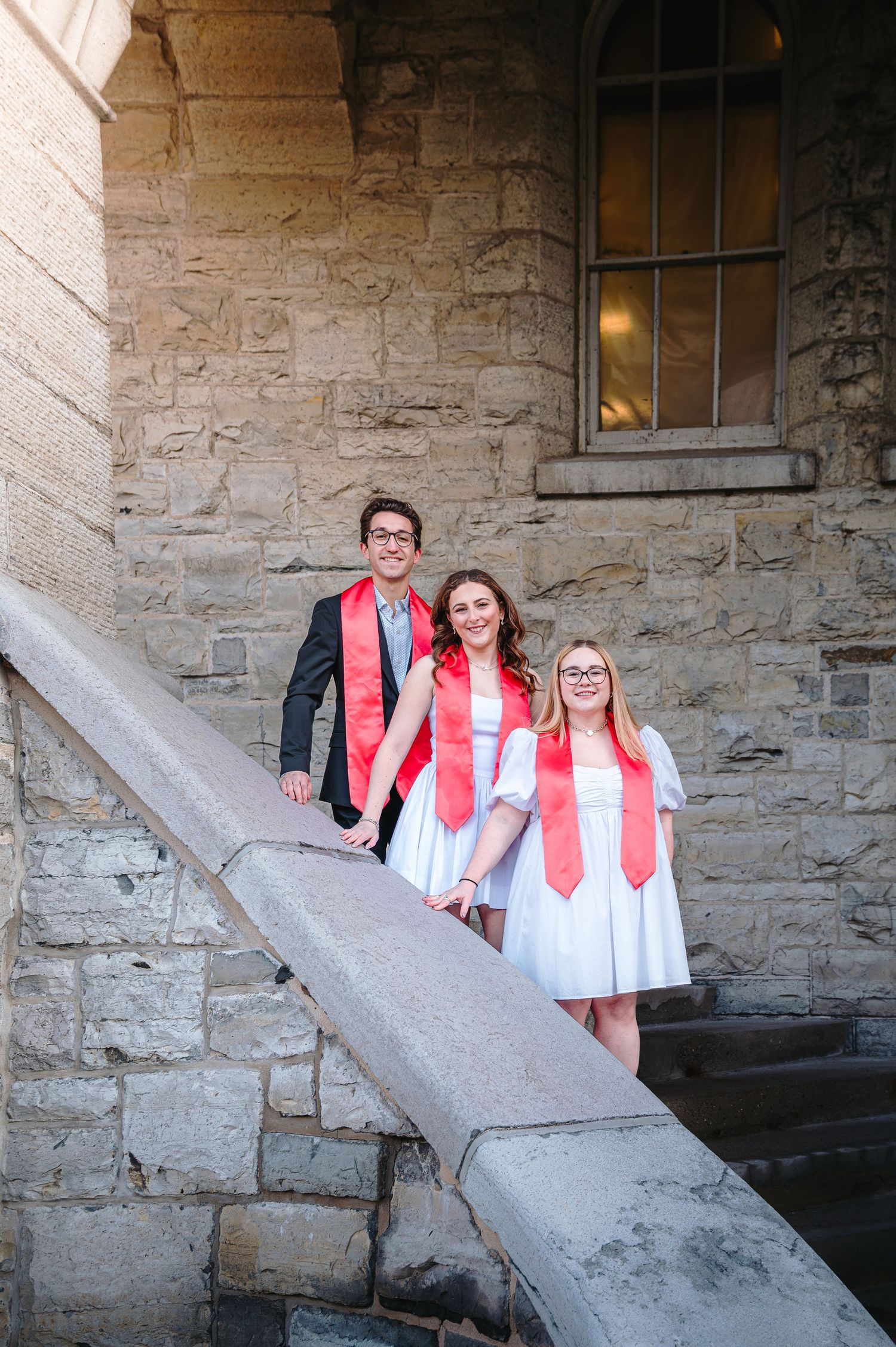 Three graduates in white dresses and red stoles pose together on stone university building steps.