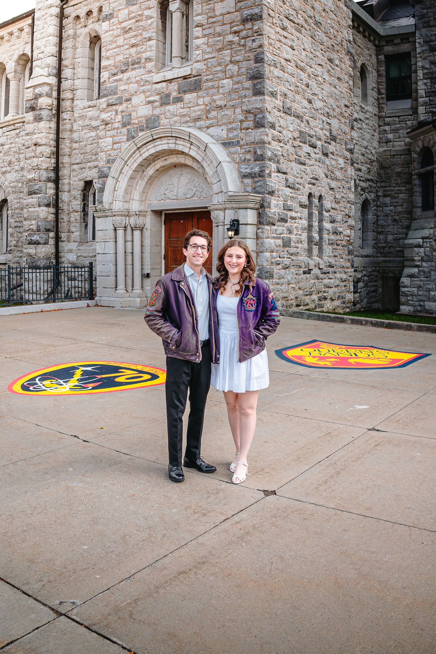 Two students in purple varsity jackets pose outside a stone university building with school logos on the ground.