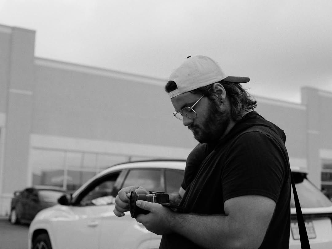 Male photographer in black t-shirt and snapback cap reviewing camera settings in parking lot.