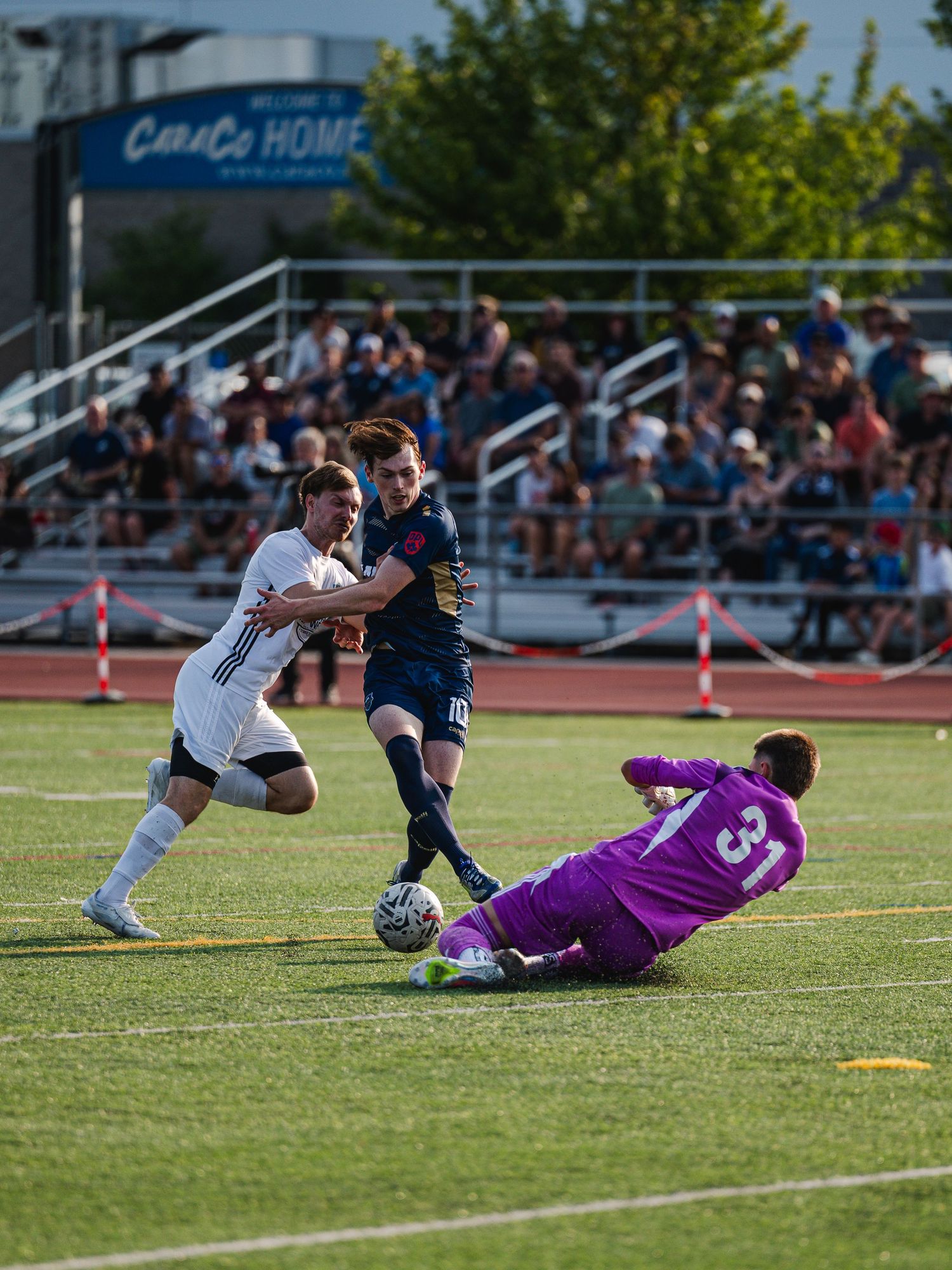 Soccer player in navy kit dribbles past goalkeeper in purple jersey during a competitive match.