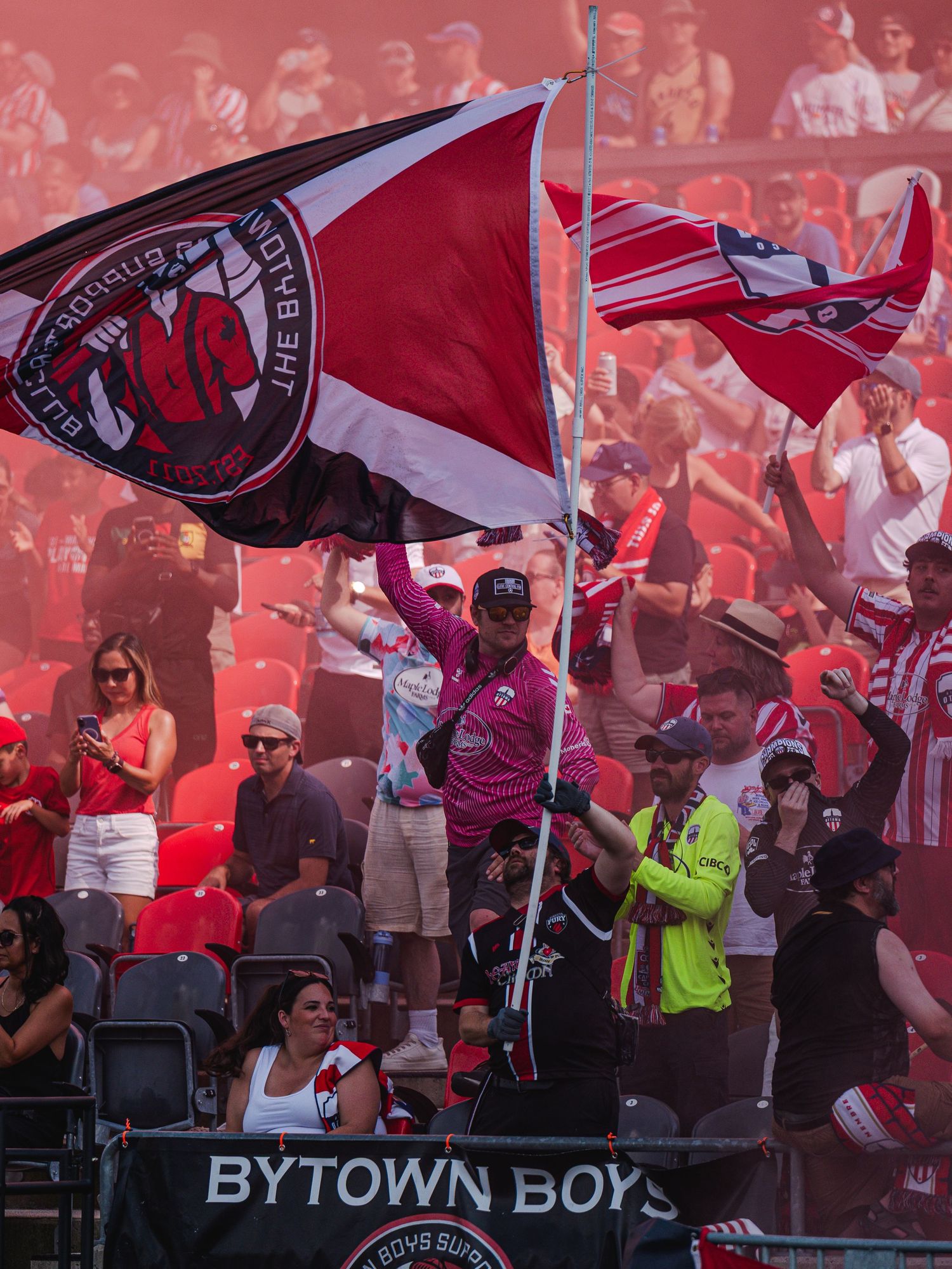 Passionate soccer fan waves large team flag amid red smoke in crowded Bytown Boys supporter section.