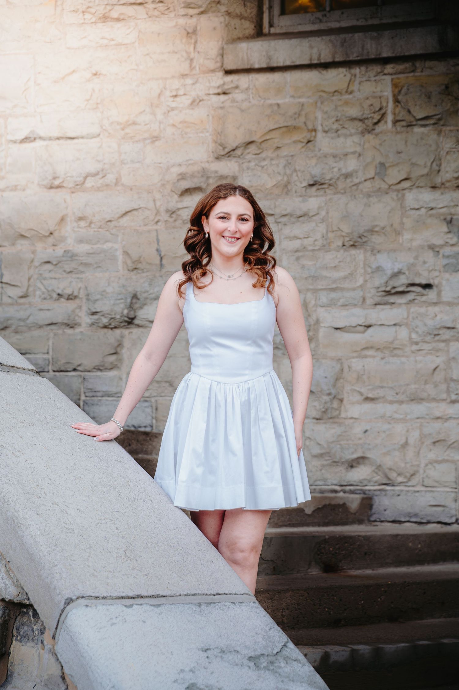 Smiling young woman in white dress poses on stone steps at a university building.