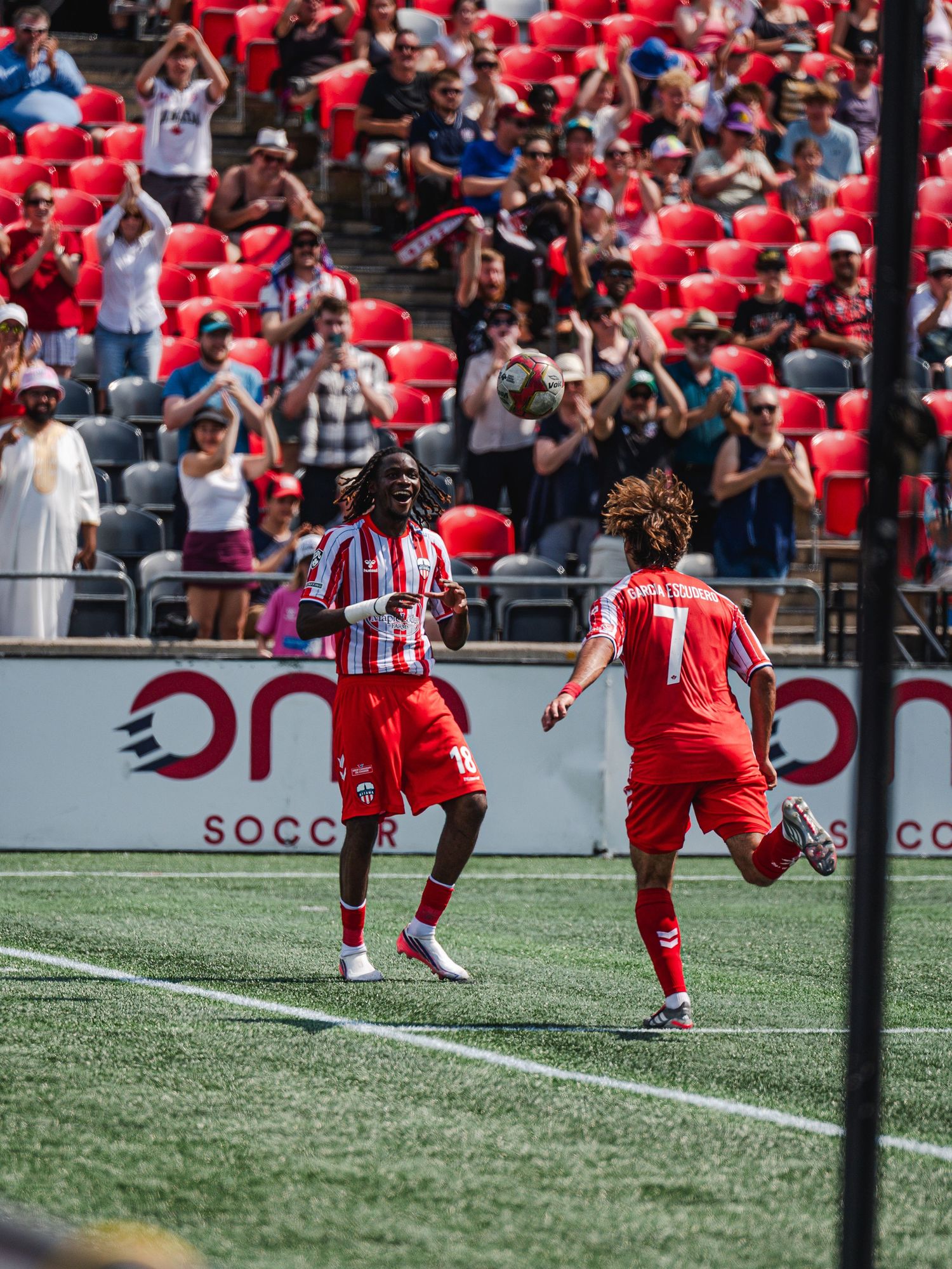 Two soccer players in red and white uniforms celebrate a goal in front of stadium crowd.