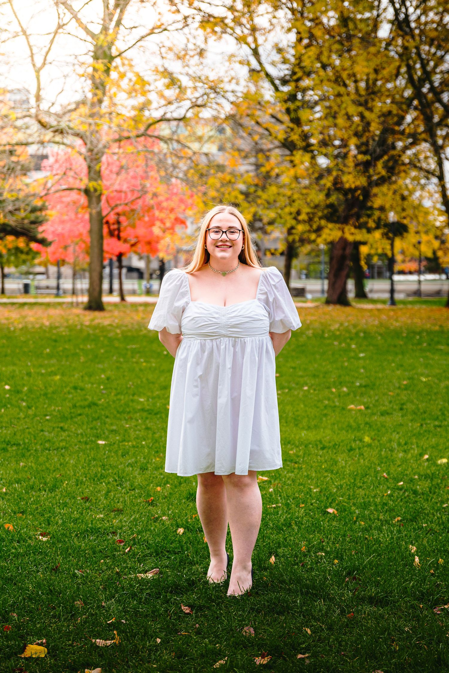 Smiling blonde woman in white dress stands barefoot on green grass with fall trees behind her.