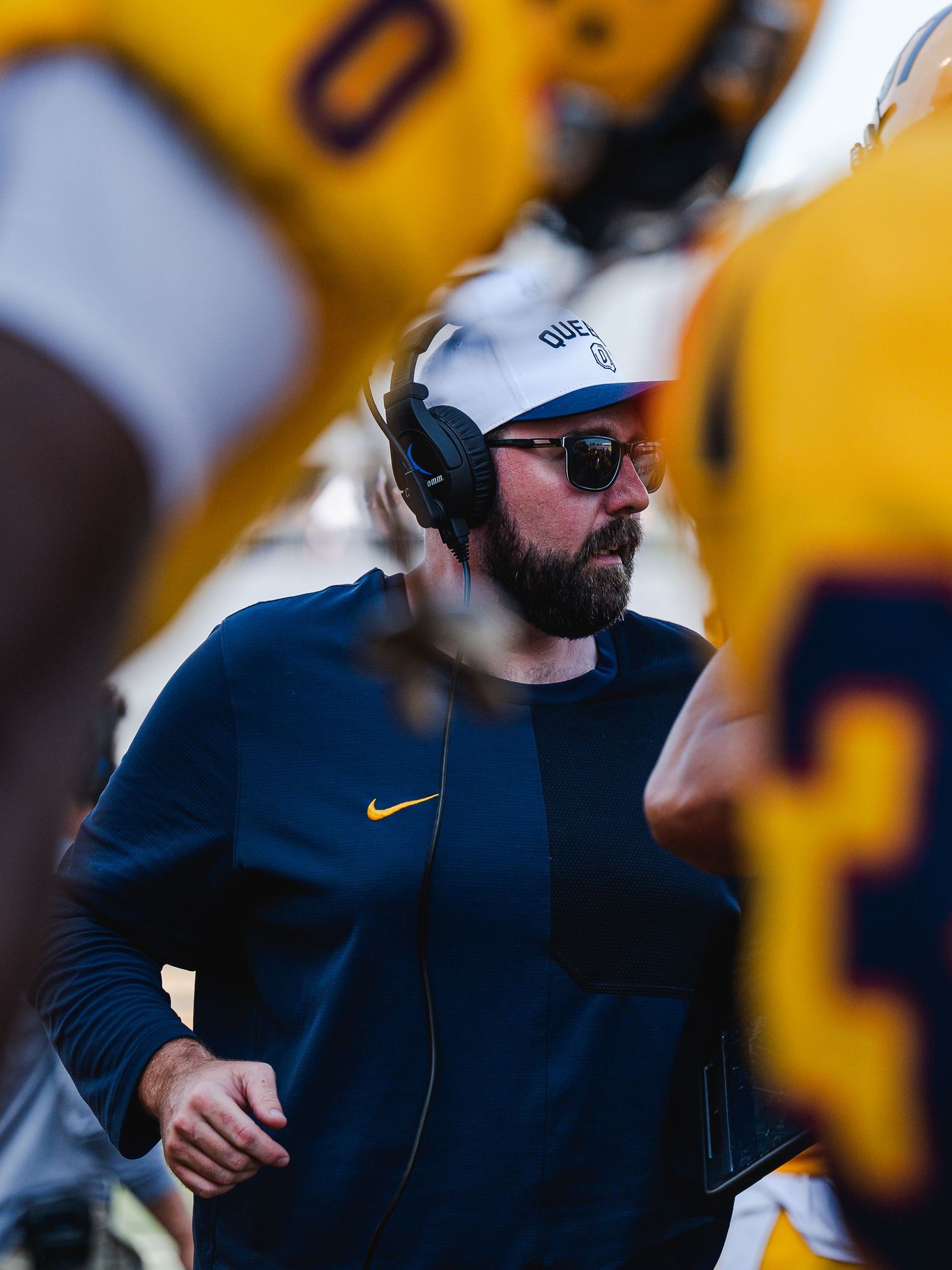 Bearded Queen's football coach wearing headset and cap observes play during college football game.