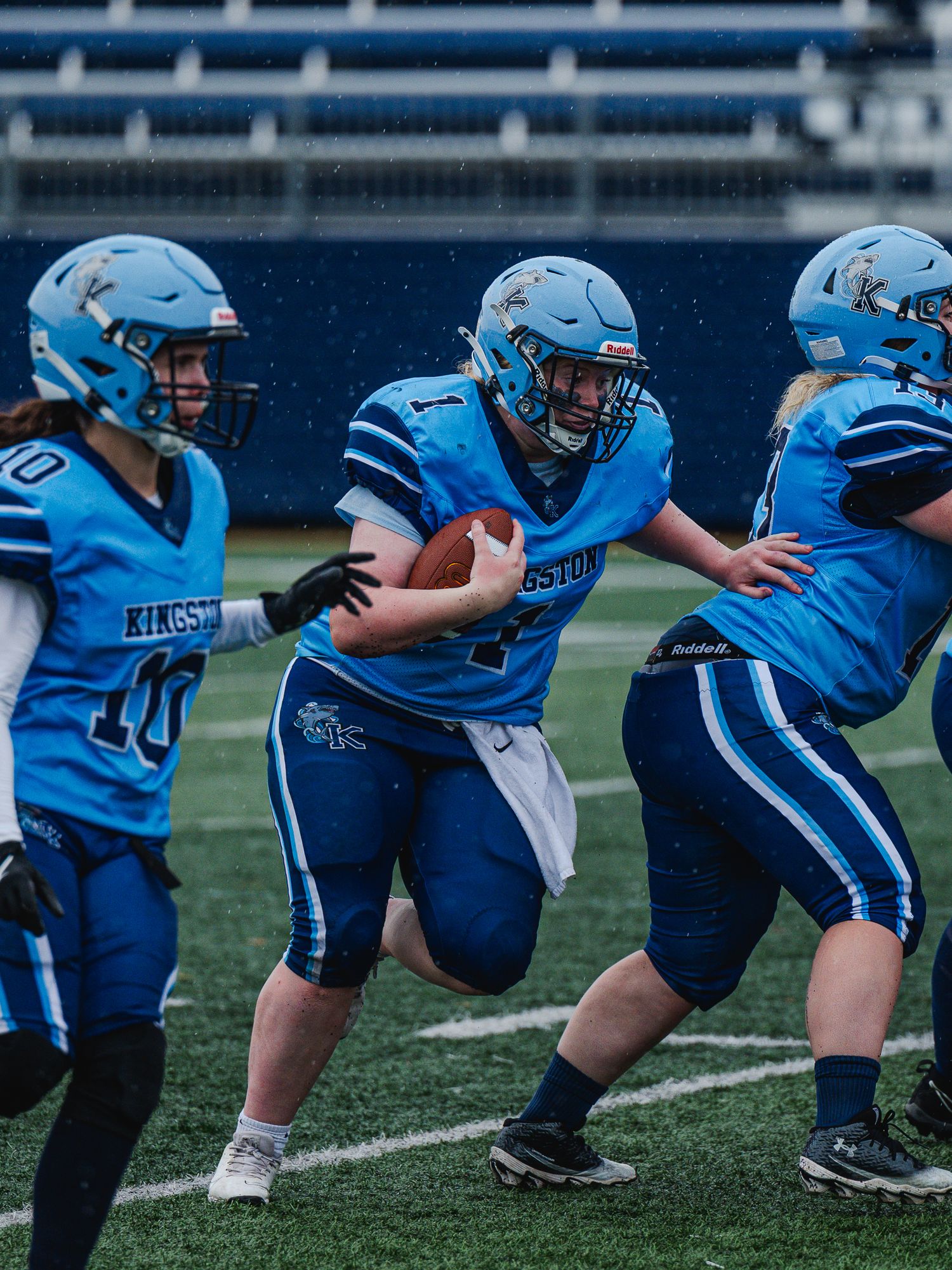 Kingston football players in blue uniforms during a rainy game, ball carrier breaks through.