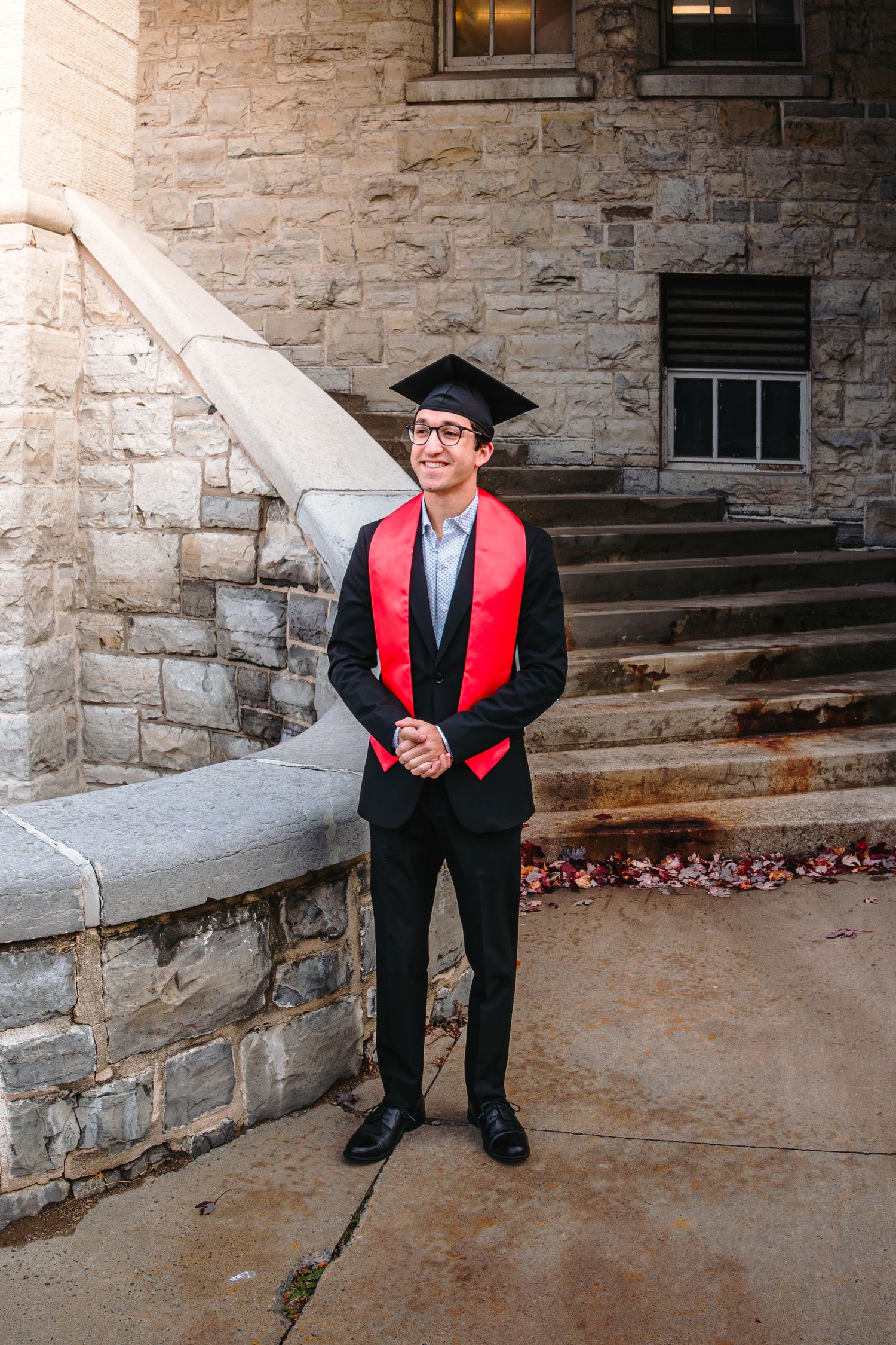 Smiling male graduate in black suit, red stole and mortarboard poses outside stone university.