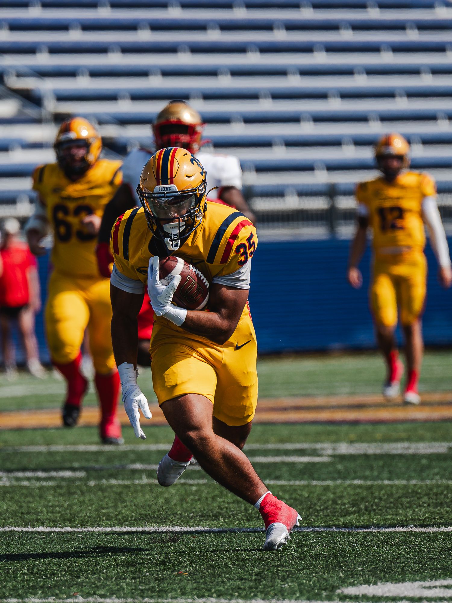 Queen's University football player in gold uniform carries ball.