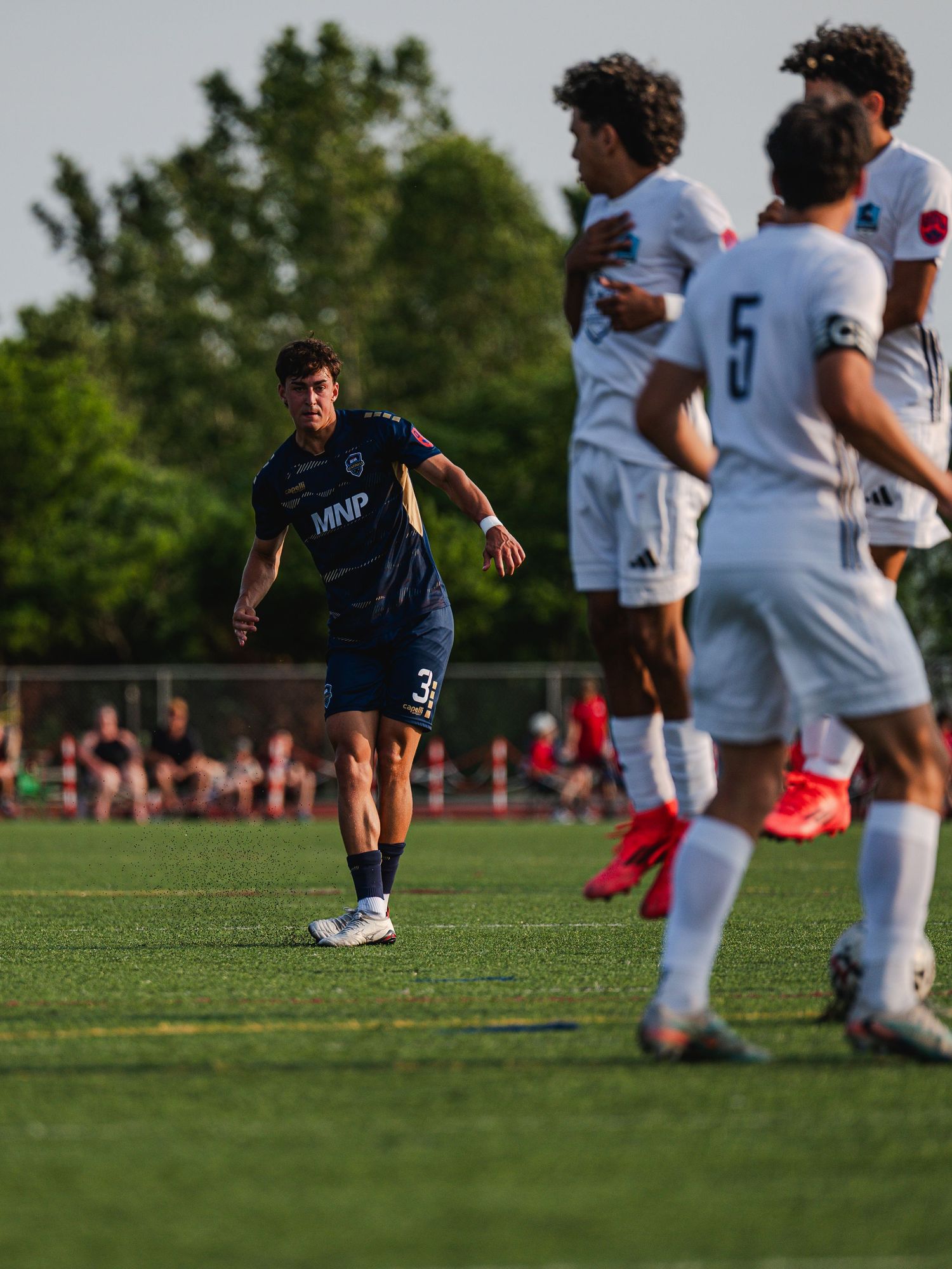 MNP player number 3 in navy kit takes a free kick toward a wall of white-uniformed opponents.
