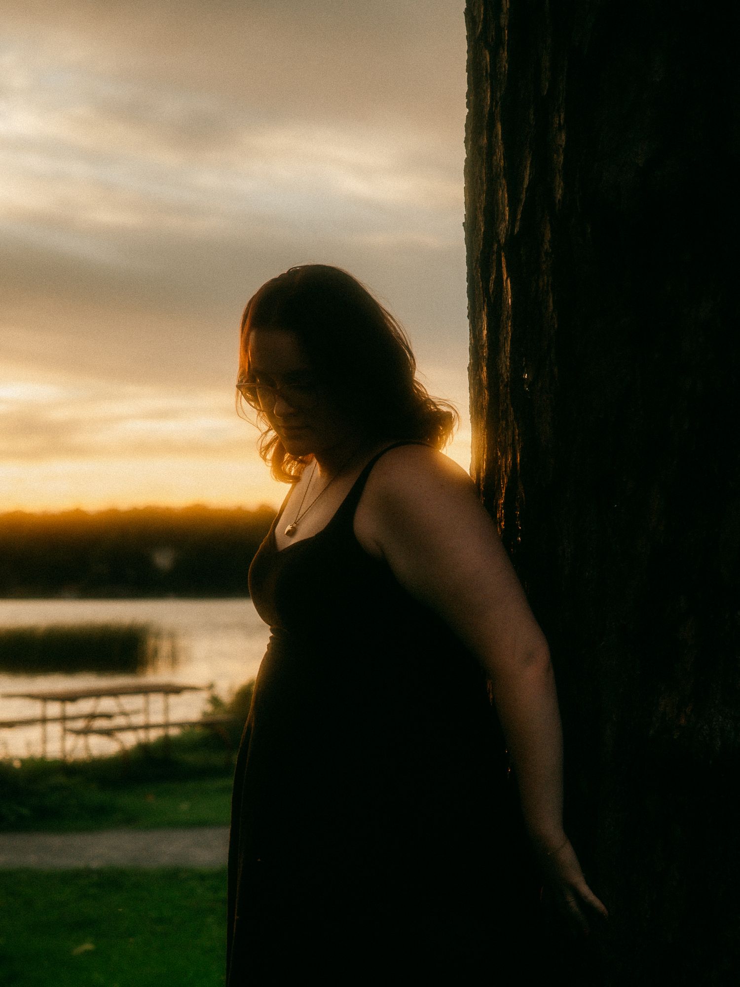 Woman in black dress leaning against tree at sunset by a lake in warm golden light.