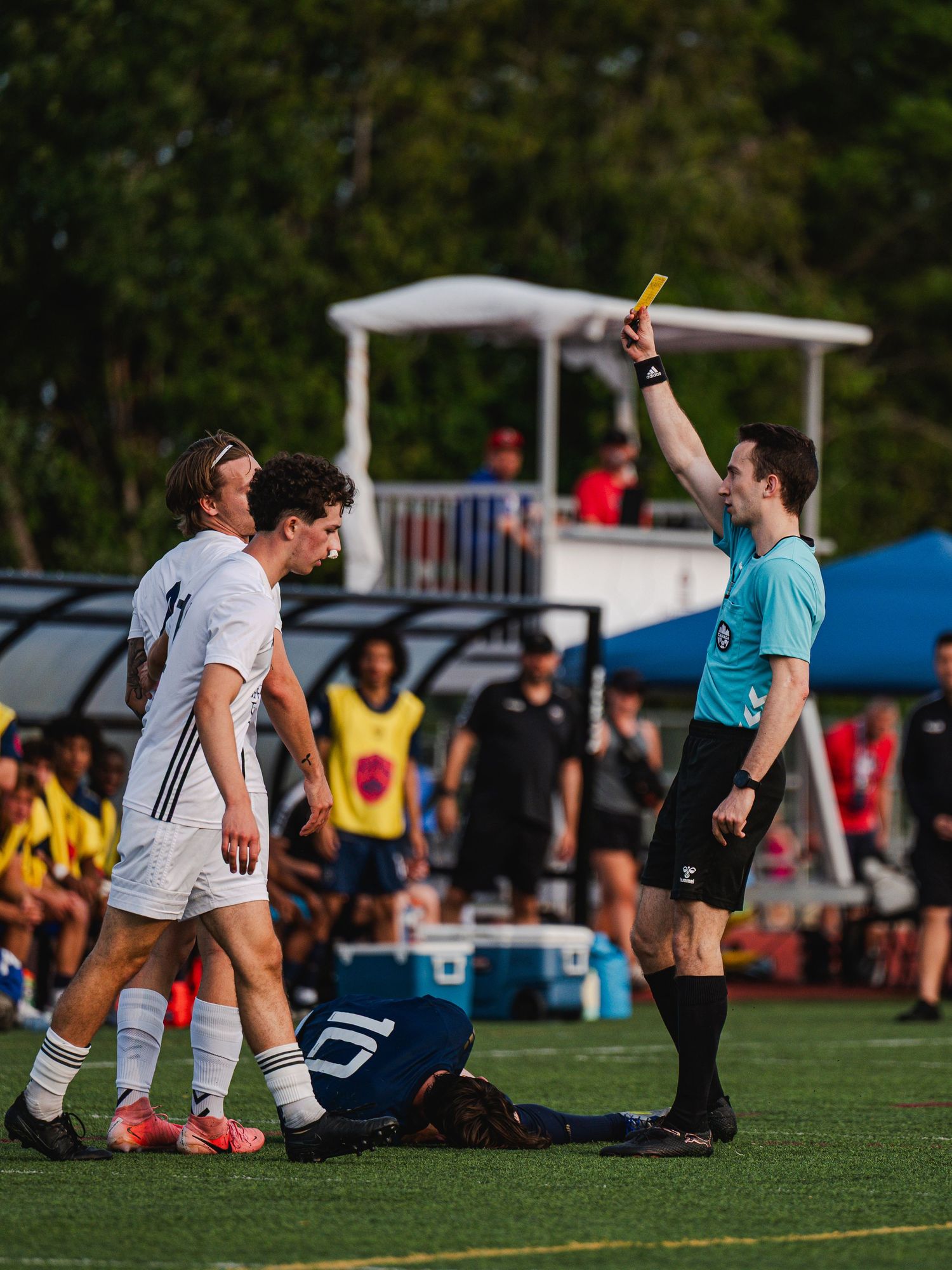 Referee in light blue shirt shows a yellow card to a white-uniformed soccer player during a match.
