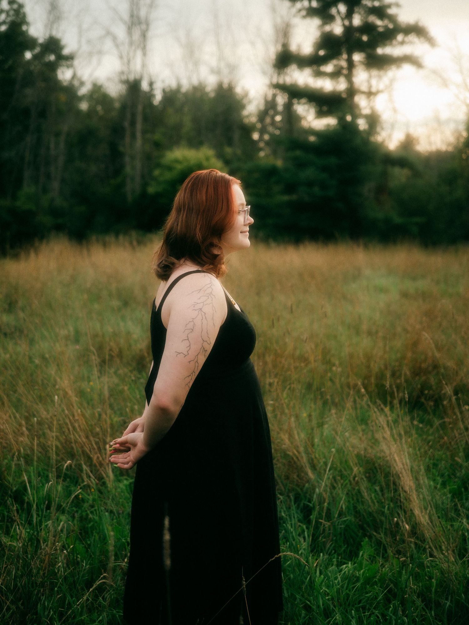 Smiling redhead woman in black dress standing in meadow with tall grass and trees at dusk.