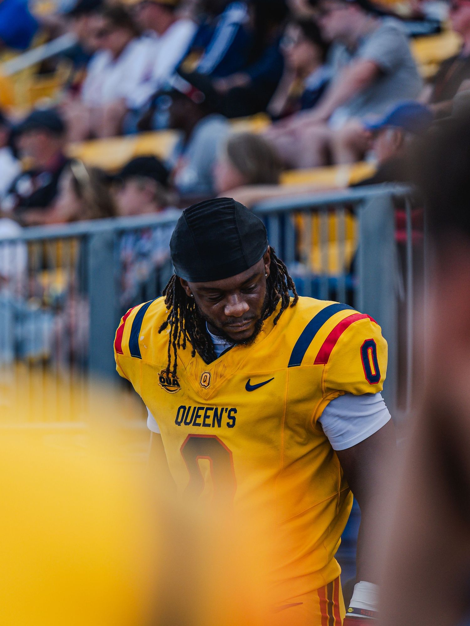 Queen's football player #0 in gold Nike uniform walks along sideline during game.