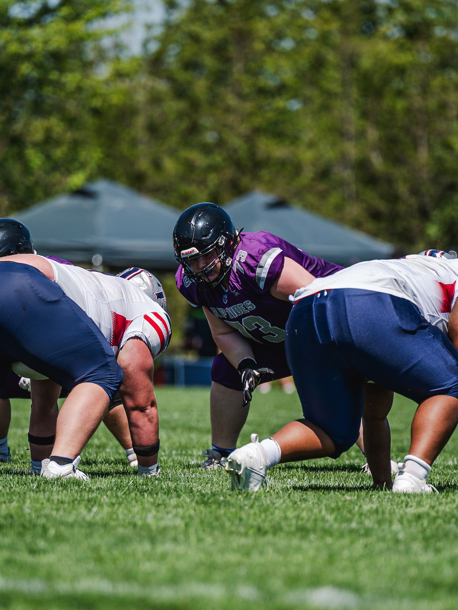 Purple team football player lines up against white-uniformed opponents on a sunny green field during an outdoor game.