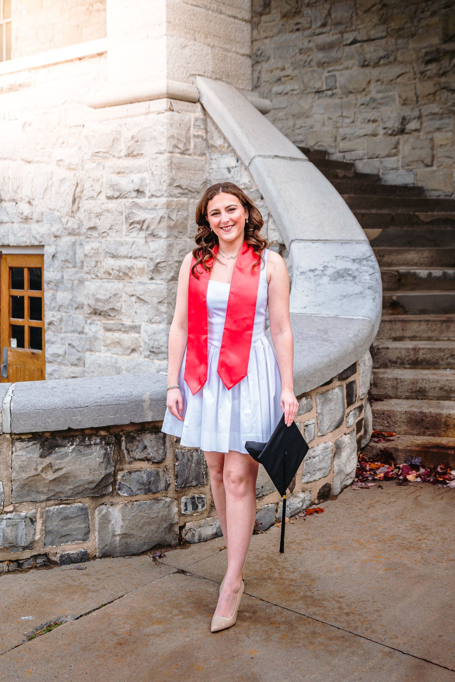 Graduating woman in white dress and red stole holds cap while standing outside stone building.