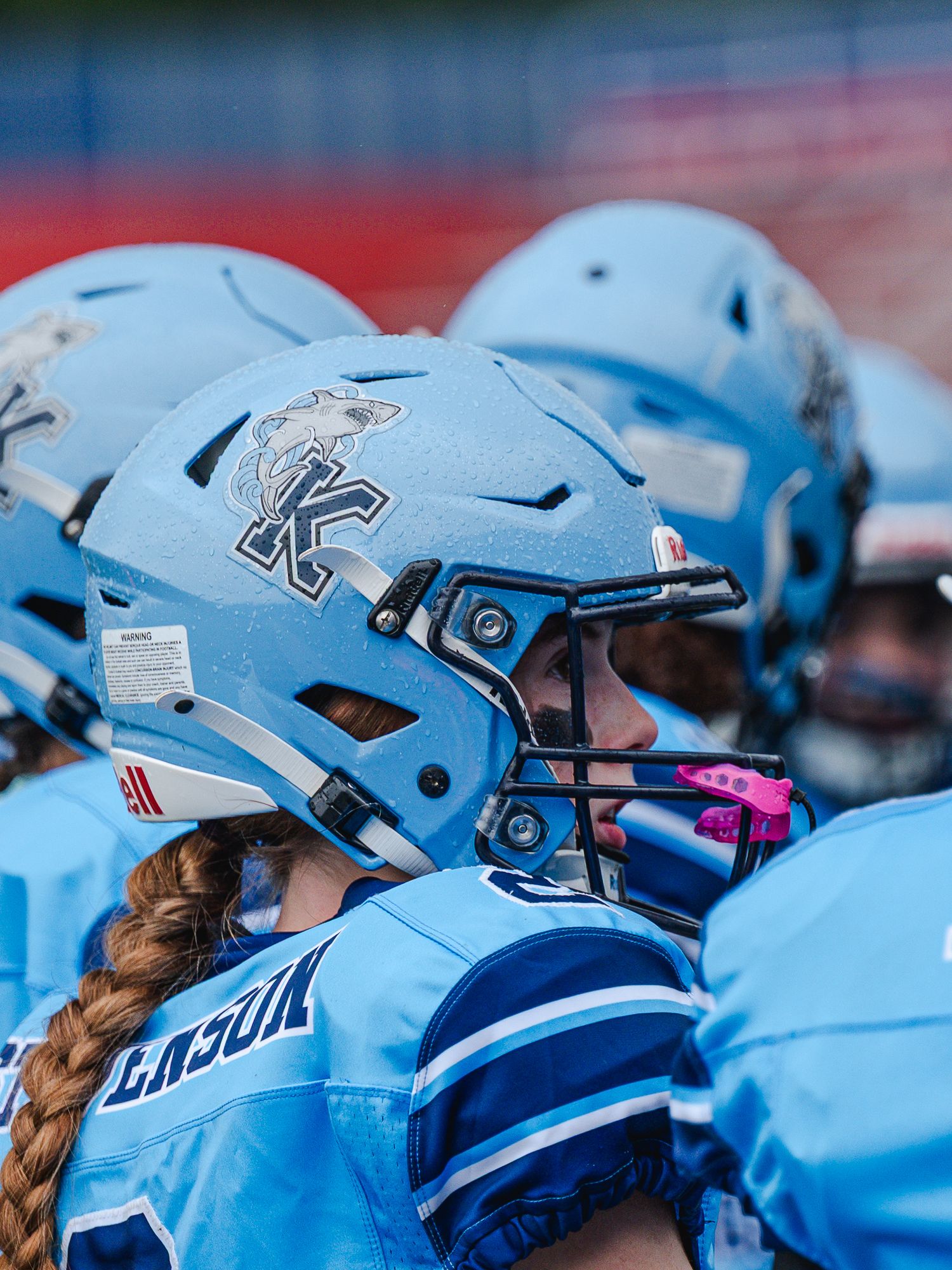 Group of female football players wearing light blue Kingston helmets with braided hair visible on the sideline.