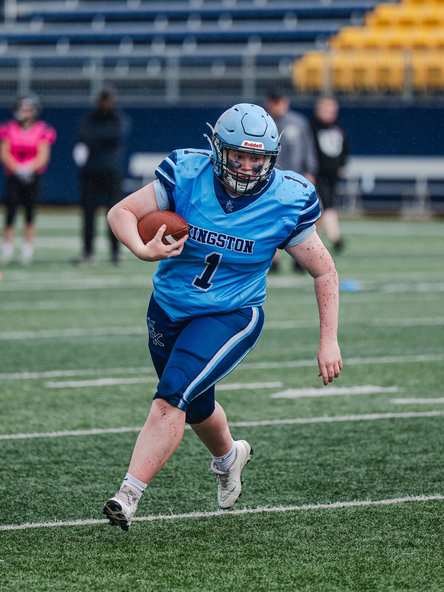 Football player number 1 in a light blue Kingston uniform carries the ball during a game on a stadium field.