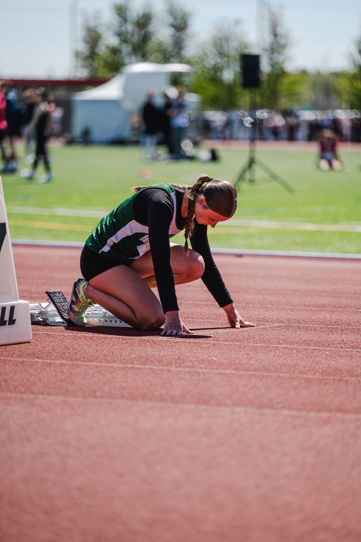 Female track athlete in green uniform positioned in starting blocks at sunny outdoor meet.