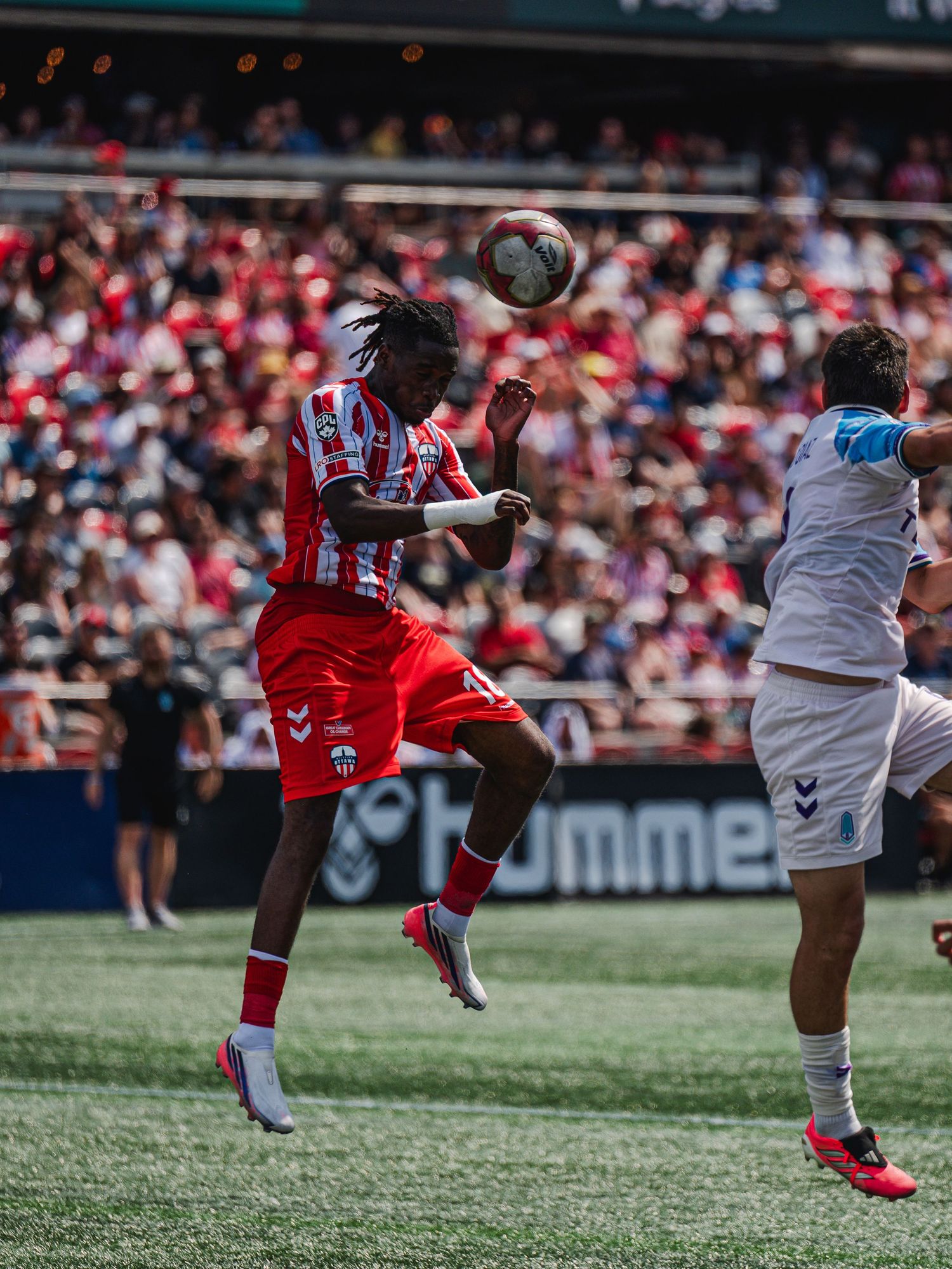 Soccer player in red and white kit wins aerial header challenge during sunny stadium match.