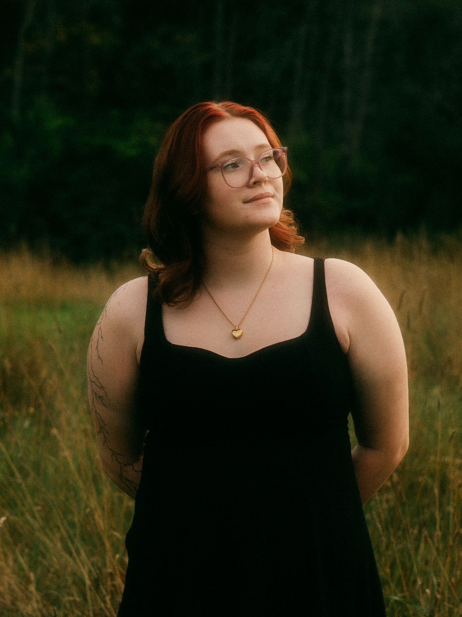 Smiling redhead woman with glasses and tattoos wearing black dress outdoors in golden meadow.