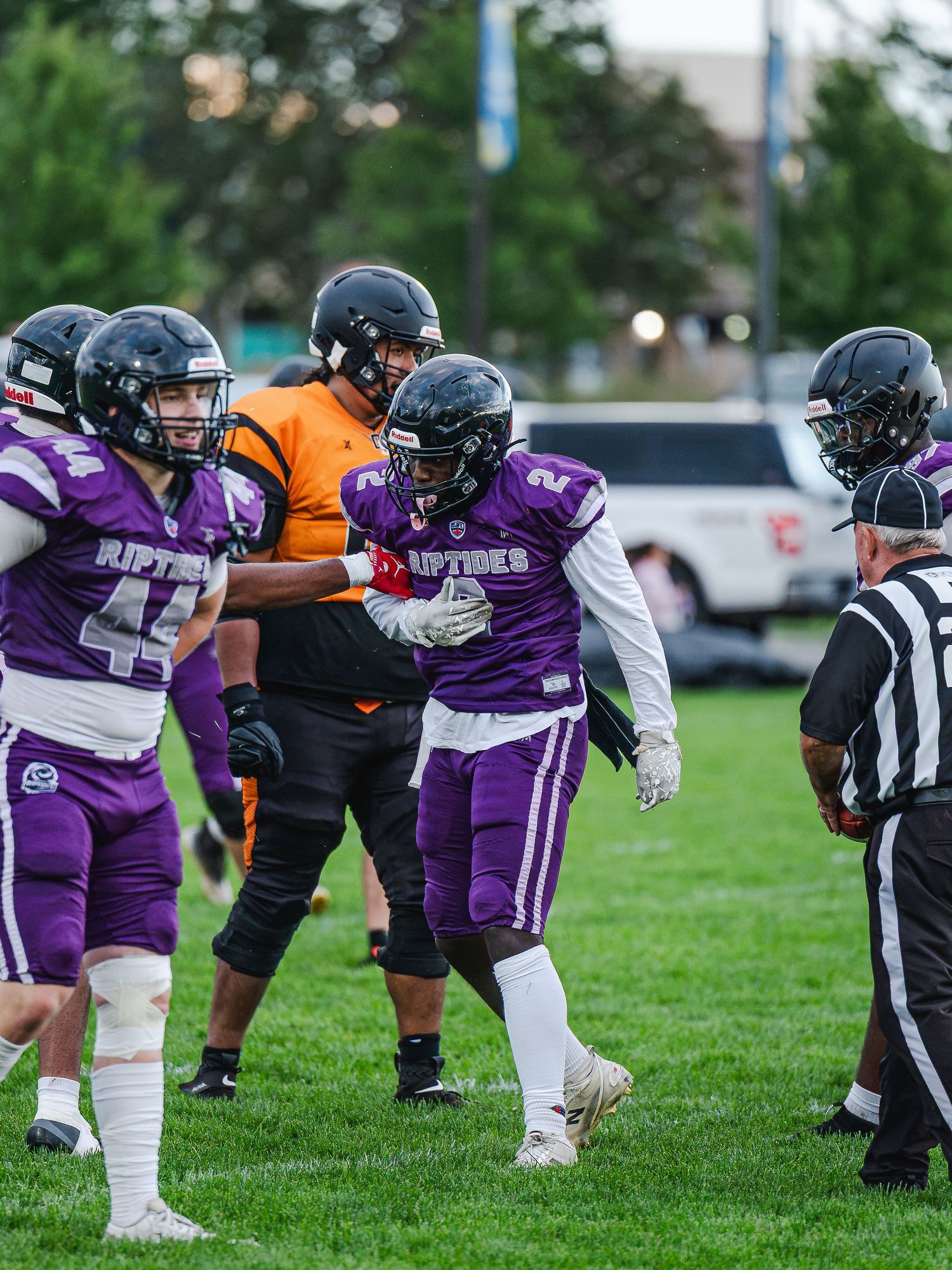 Riptides football players in purple jerseys interact with referee during game on green field.