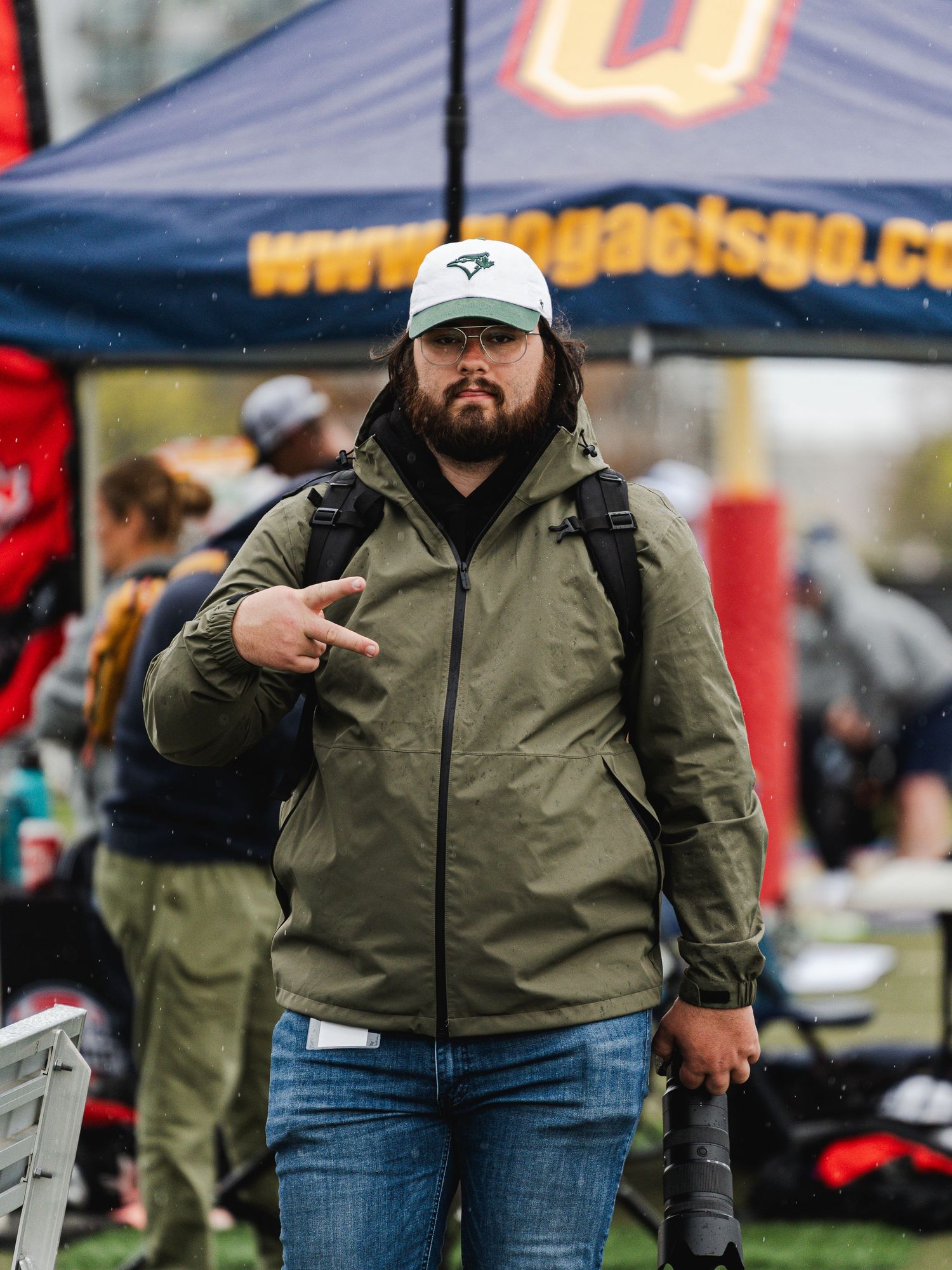 Male photographer in olive jacket and Blue Jays cap pointing at camera at outdoor sports event.