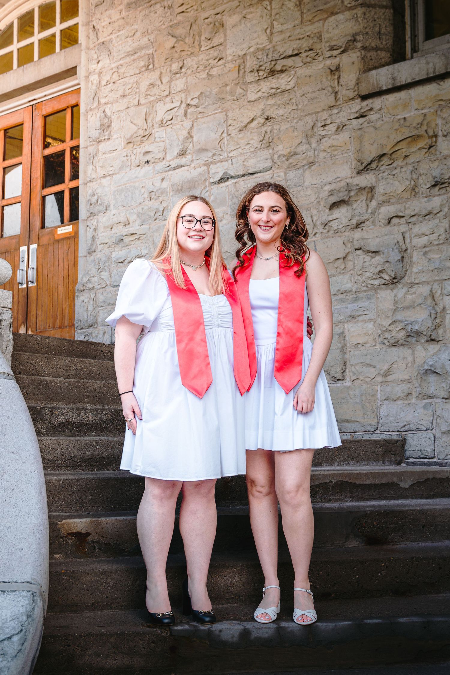 Two female graduates in white dresses and red stoles smile on stone university building steps.
