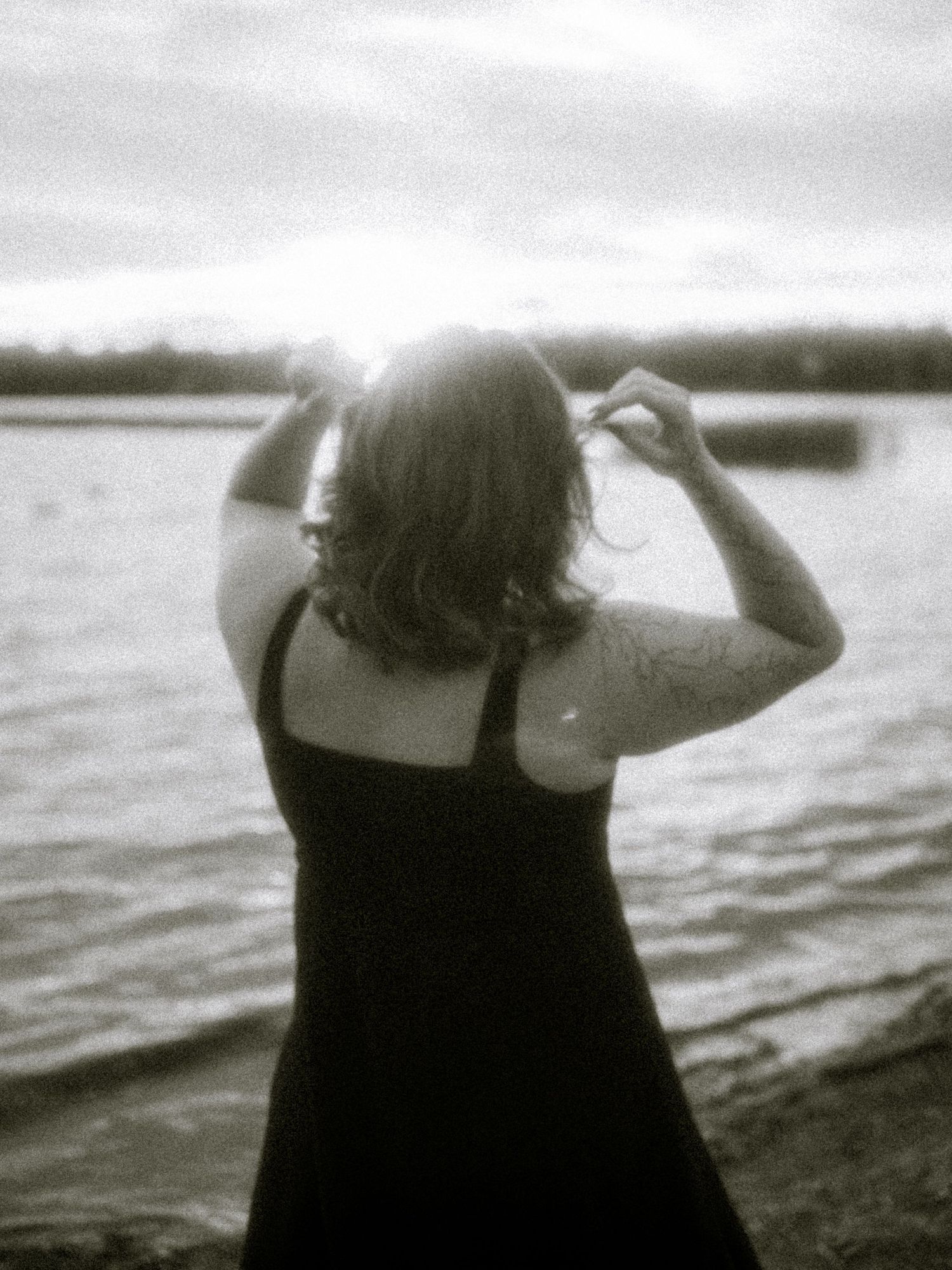Black and white photo of a woman in a dark dress standing by a lake, hands raised to her hair, sun glowing behind her.