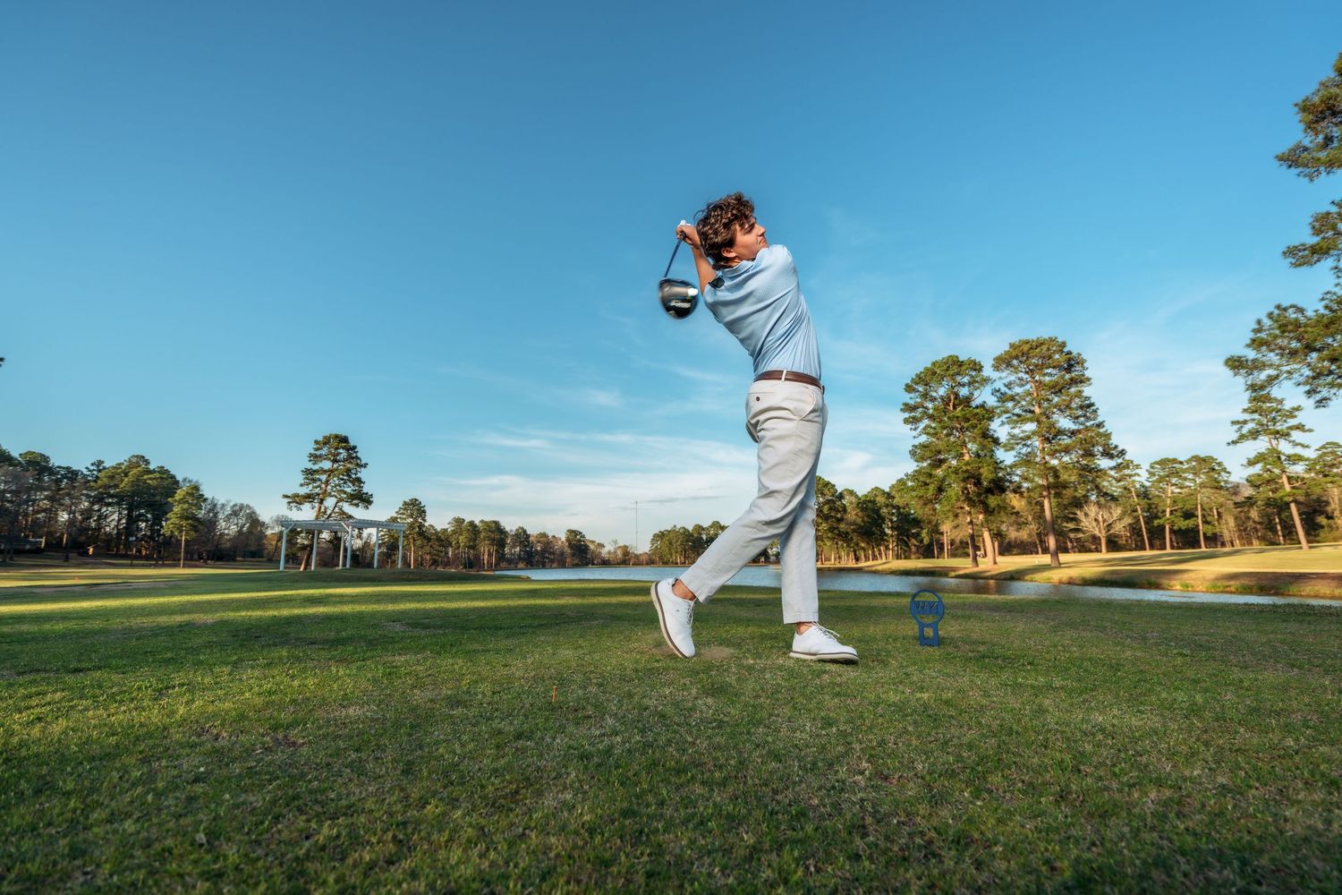 Golfer completing a full swing on a lush golf course surrounded by pine trees under a clear blue sky.