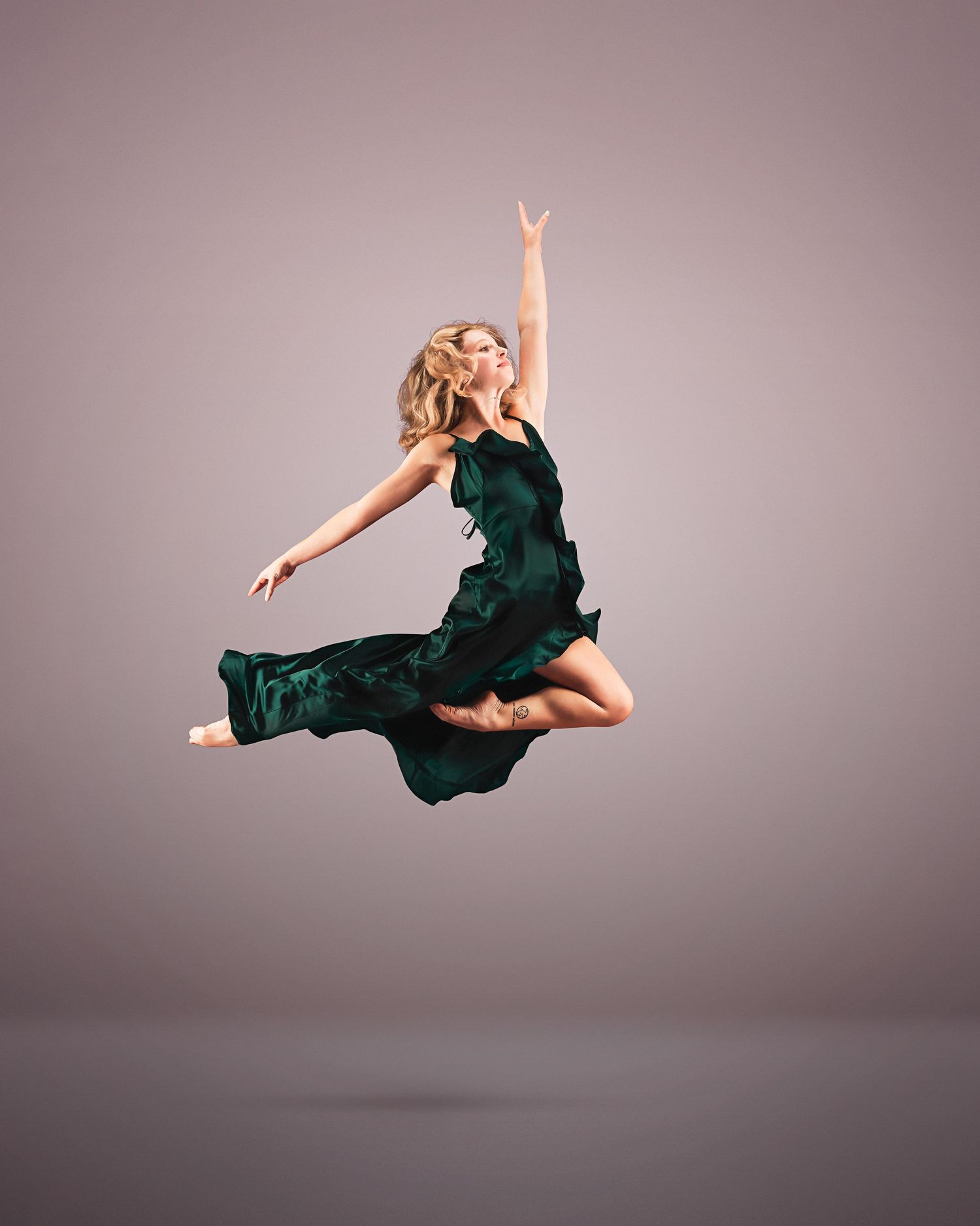 Dancer in a green dress leaps gracefully mid-air against a neutral background, arms raised in an elegant pose.