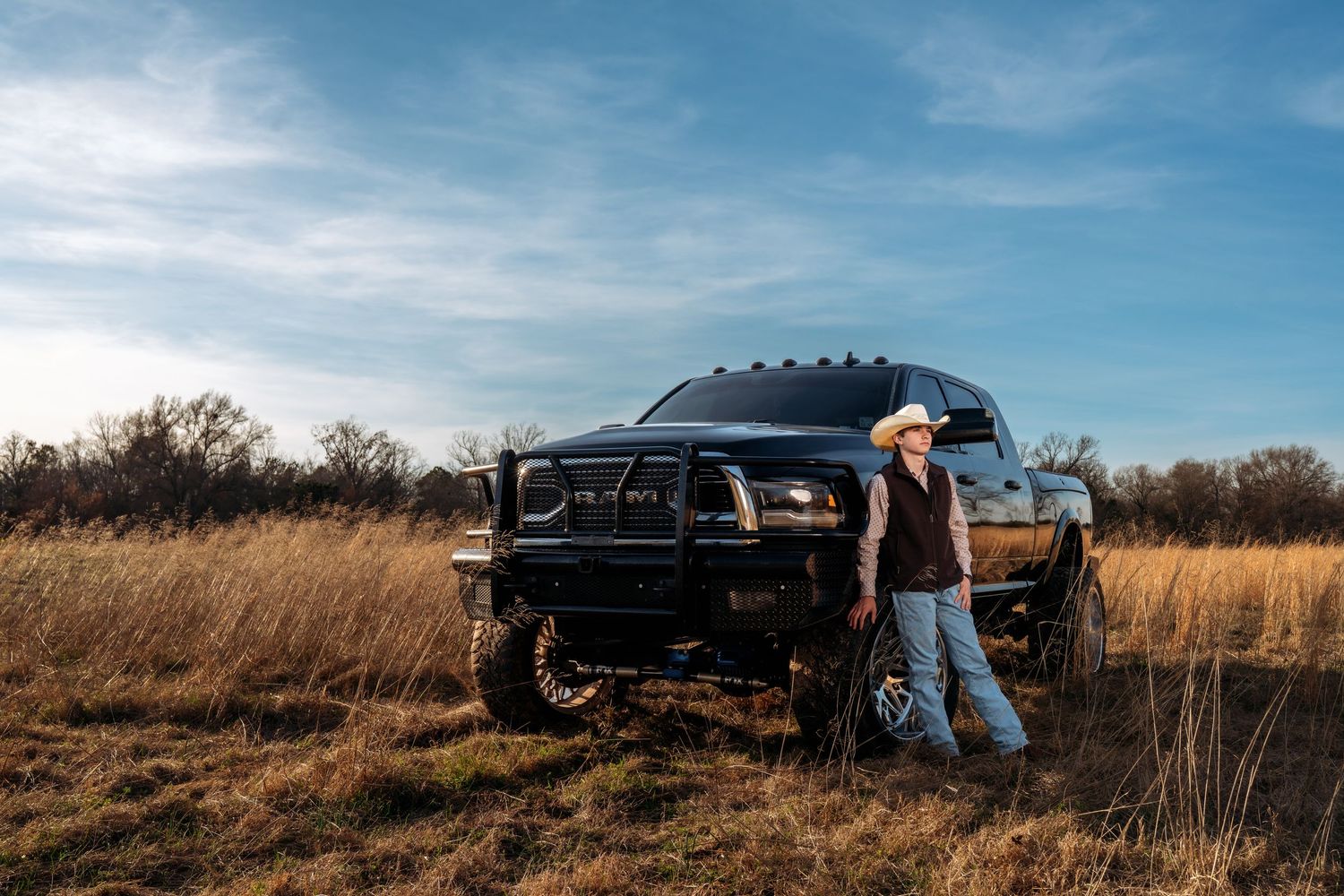 A man in a cowboy hat leans against a lifted black Ram truck in a golden field under a blue sky.