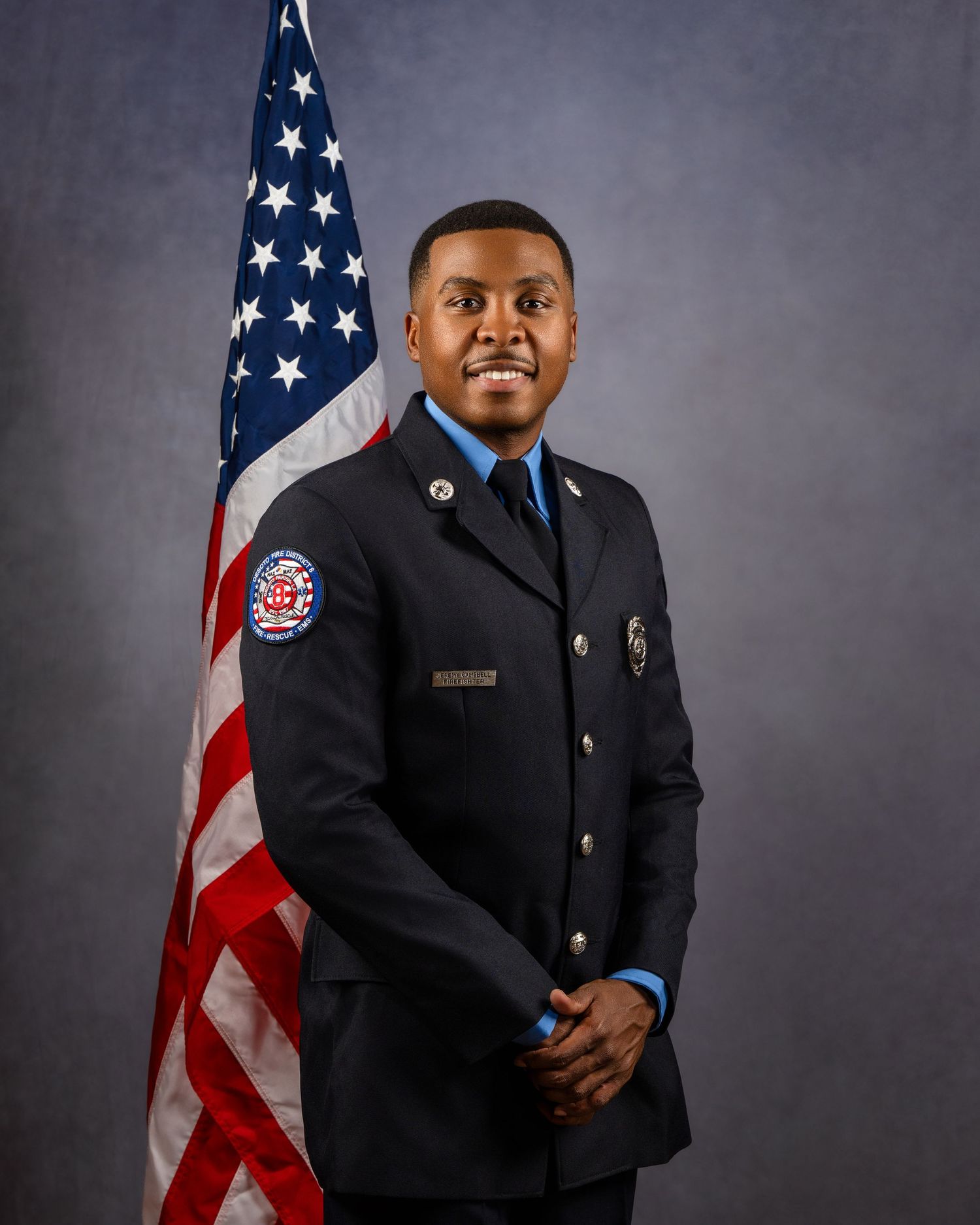 Firefighter in dress uniform smiling proudly before an American flag in an official portrait photo.