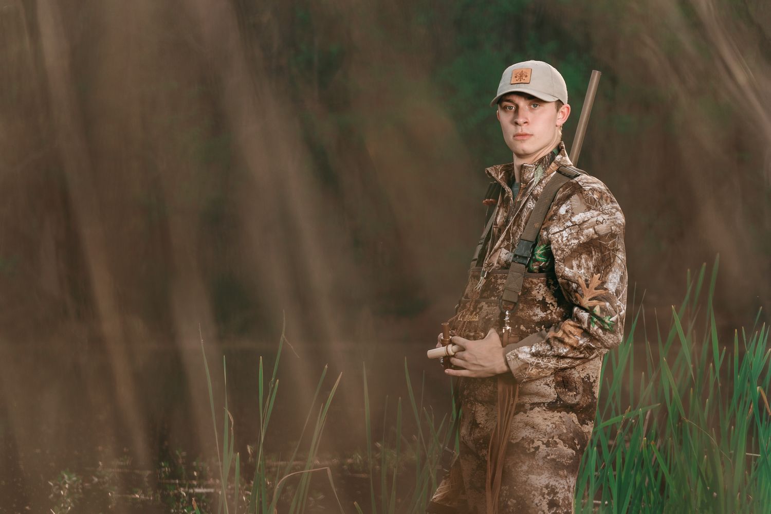 Young man in camouflage hunting gear standing in tall grass holding a rifle.
